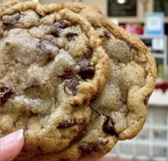 Close-up of a large chocolate chip cookie held by a person's hand, with a store background visible behind.