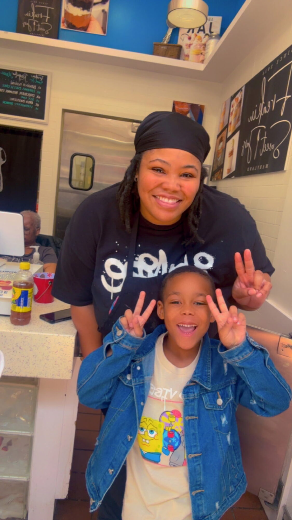 Smiling woman and child making peace signs indoors, background with chalkboard and posters, woman wearing headscarf, child in denim jacket.