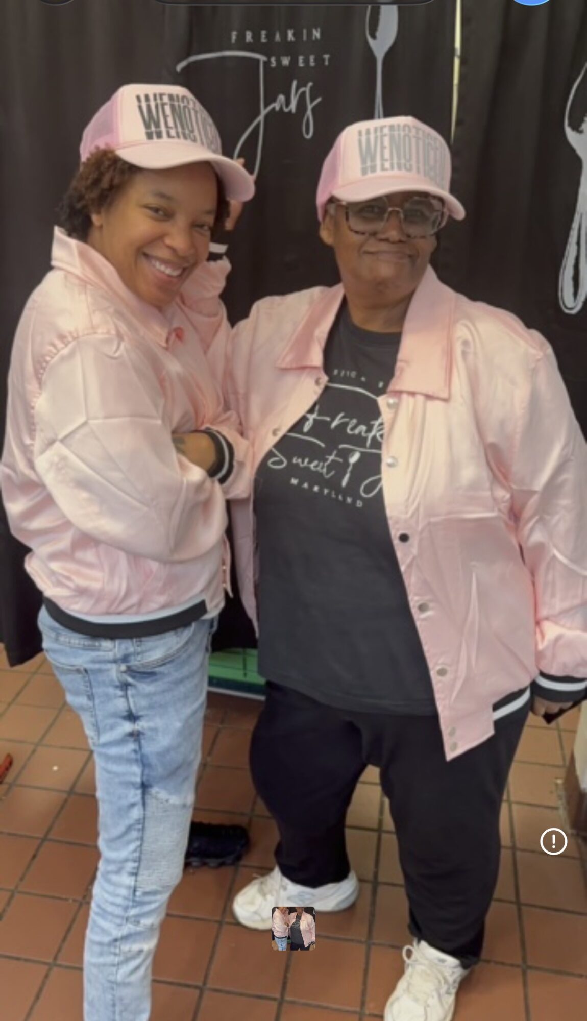 Two women wearing pink jackets and pink hats with white text, standing together and smiling.