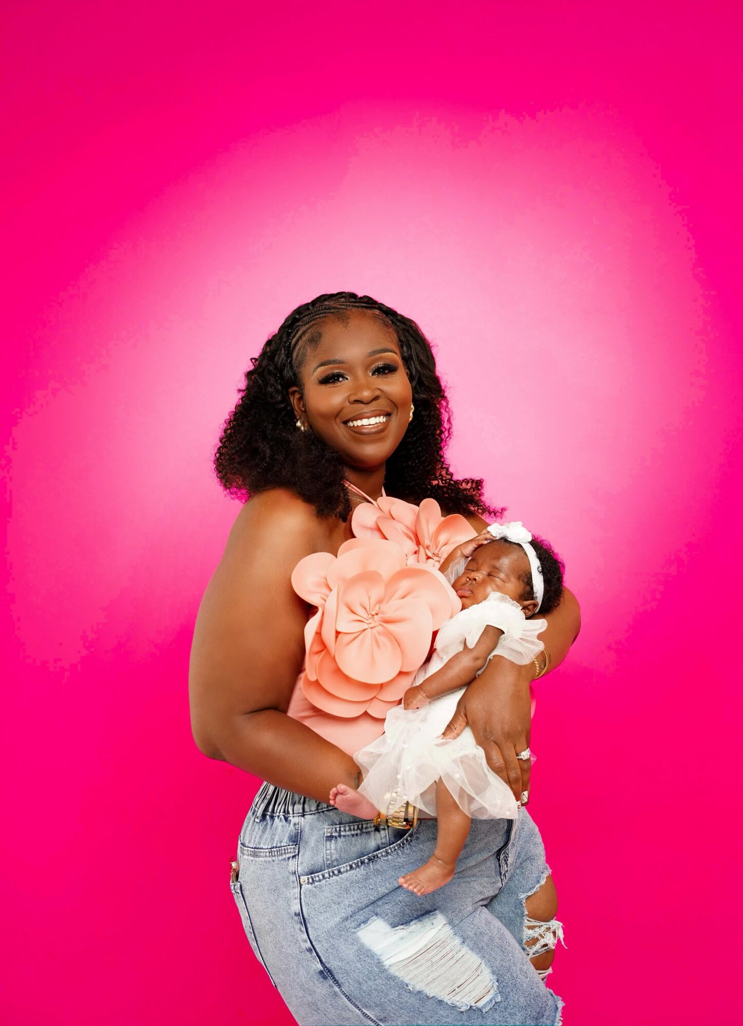 Smiling woman holding a baby against a pink background, wearing a floral top and ripped jeans.
