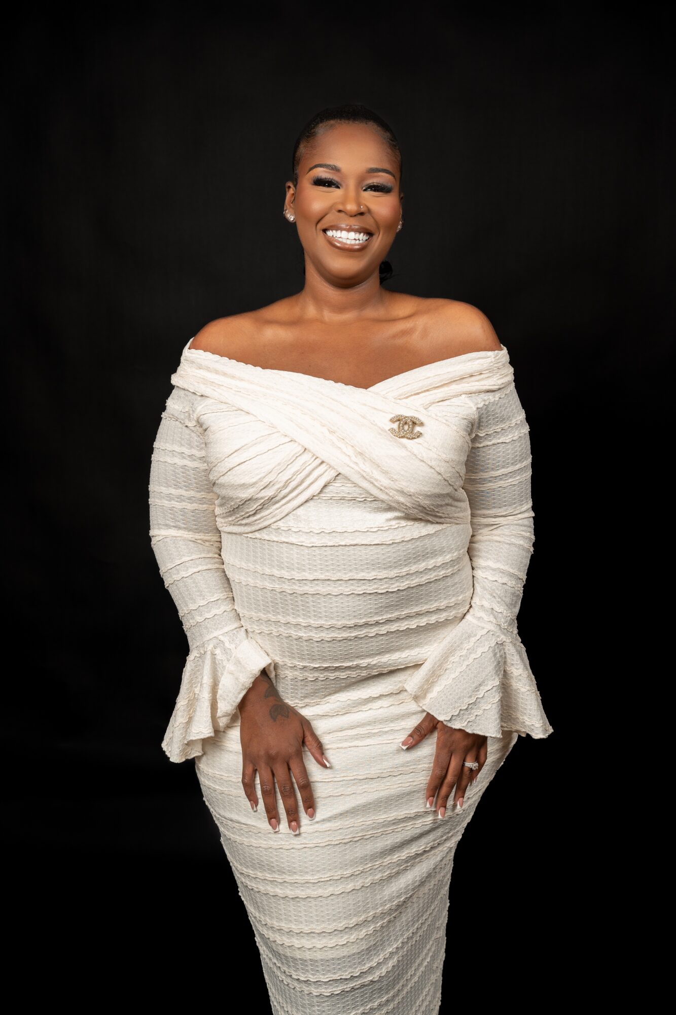 Smiling woman in off-shoulder white dress with ruffled sleeves, standing against black background.