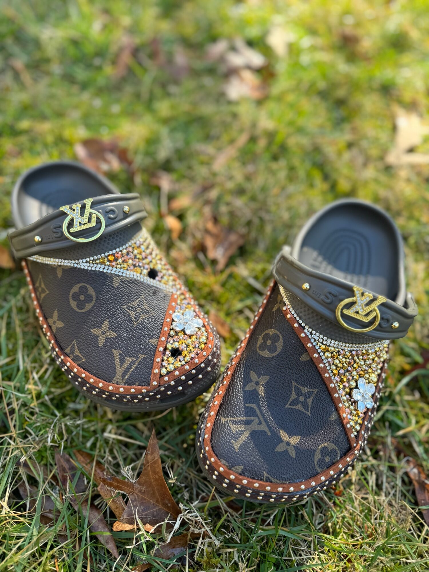 Pair of children's shoes with Louis Vuitton pattern, decorated with beads and flowers, placed on grass with fallen leaves.