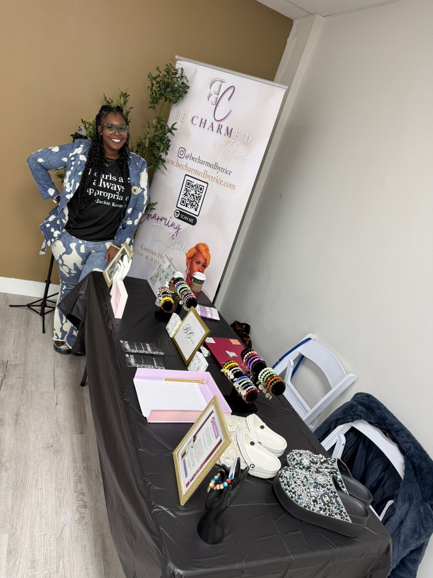 Young girl with glasses and a blue polka dot jacket stands next to a table with colorful accessories and a promotional banner.