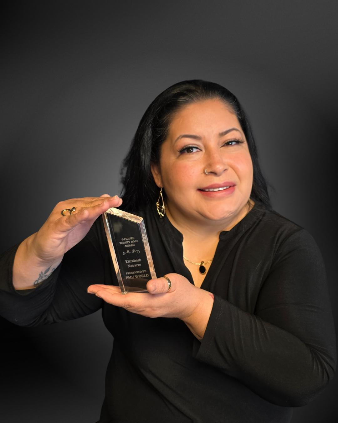 Woman holding an award plaque, smiling, wearing a black top and gold earrings, against a dark background.