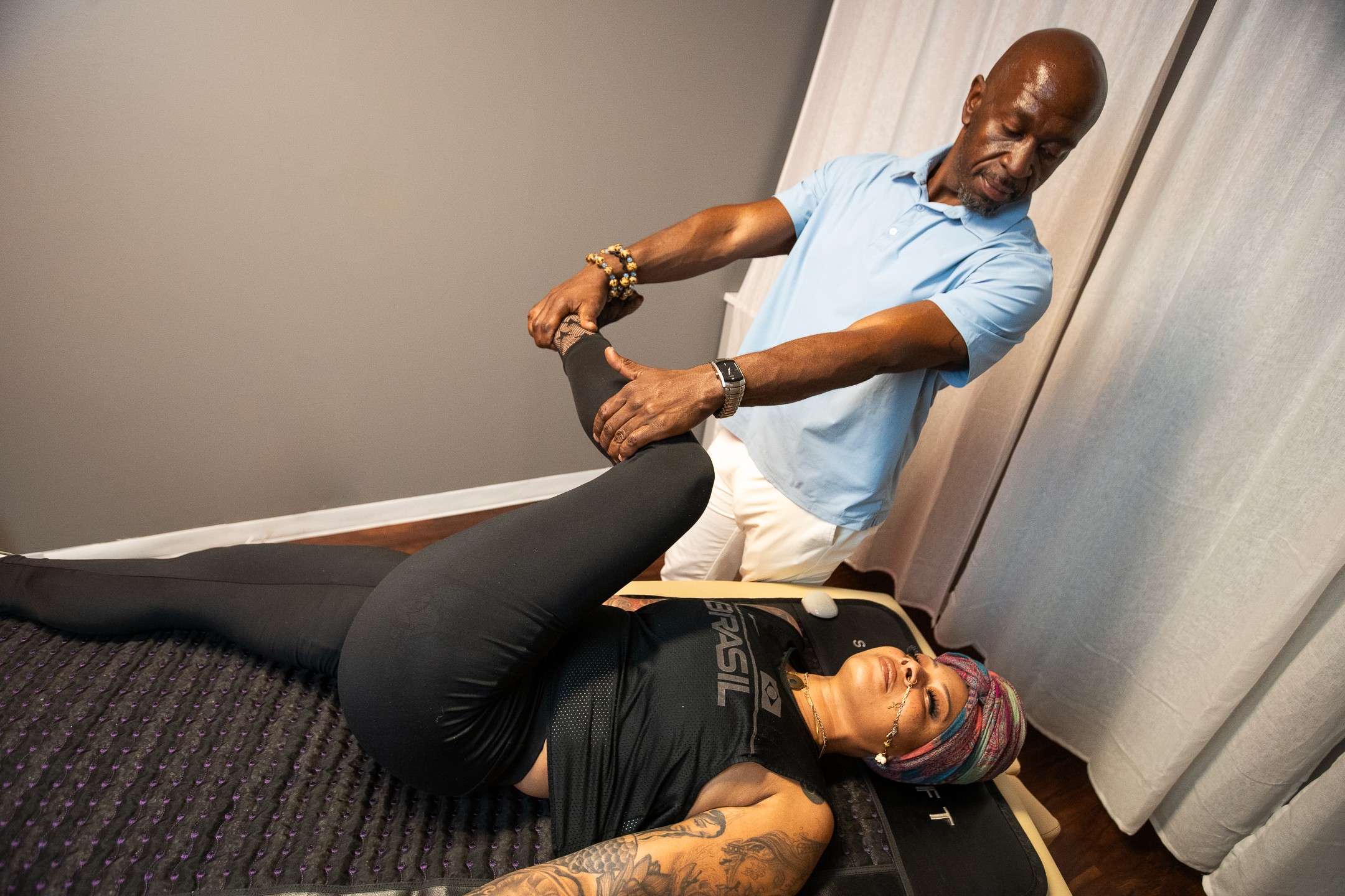 A man standing and a woman lying on the floor, holding her legs up, in a room with curtains. 20 words.