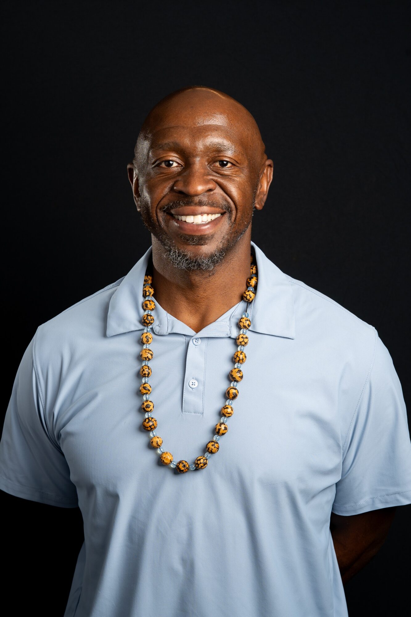 Smiling man with a beard wearing a light blue polo shirt and a beaded necklace, against a dark background.