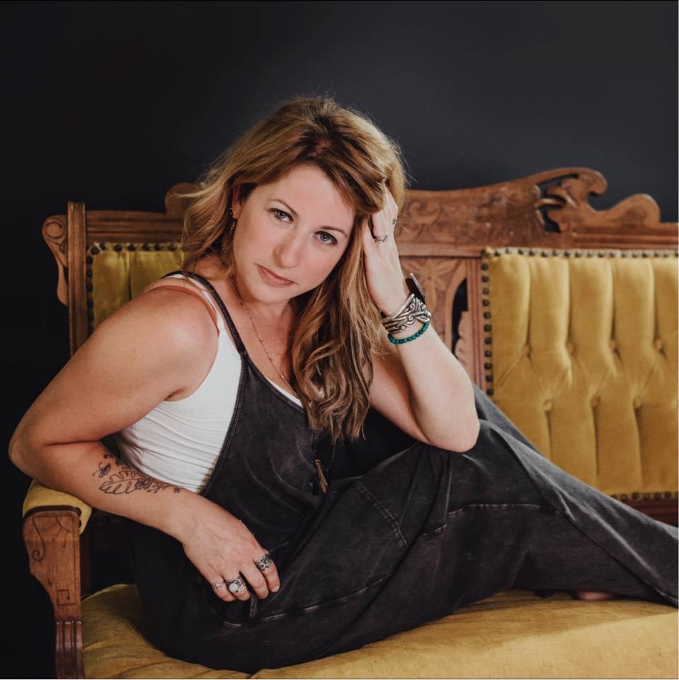 Woman with wavy hair sitting on a vintage wooden and yellow upholstered sofa, looking at the camera, with a dark background.