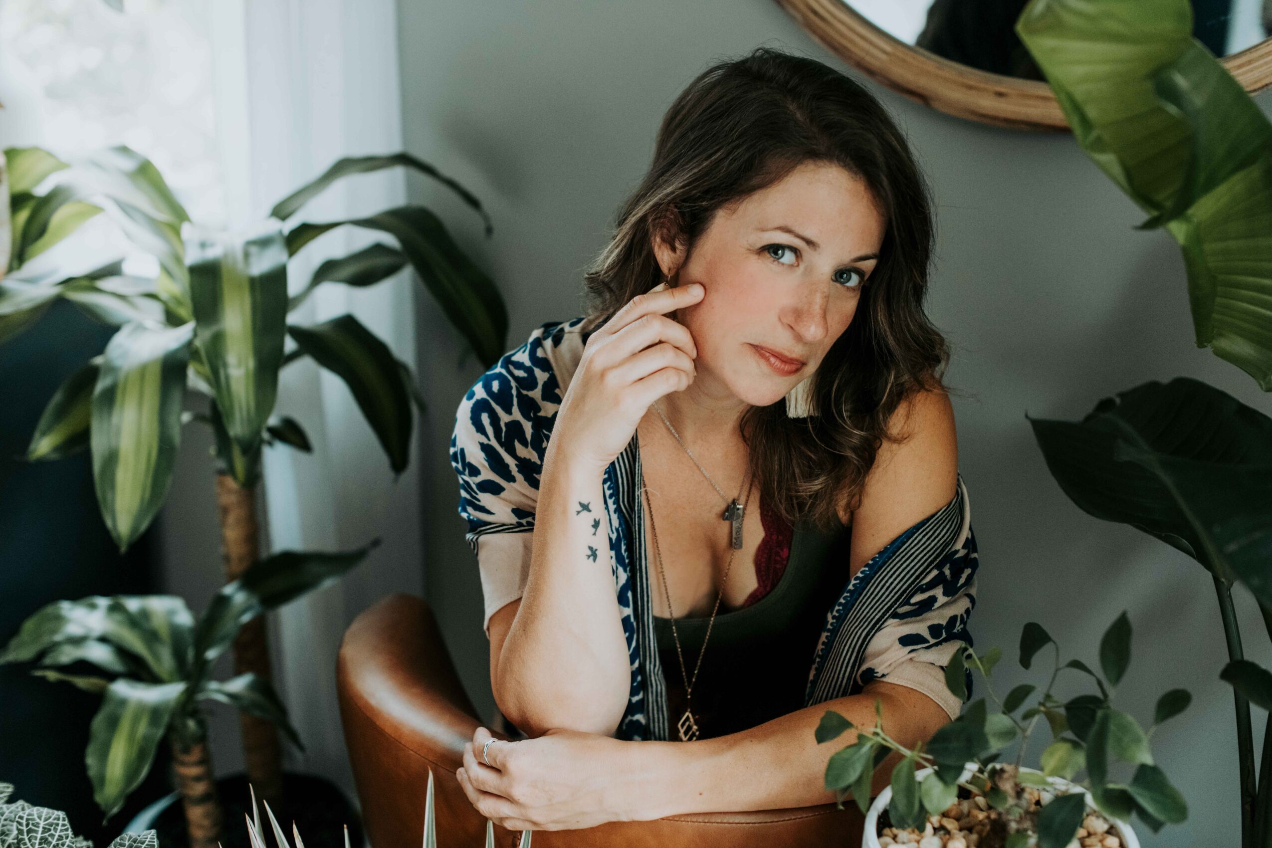 Woman with dark hair and patterned clothing sitting among green plants, looking at the camera with a thoughtful expression.
