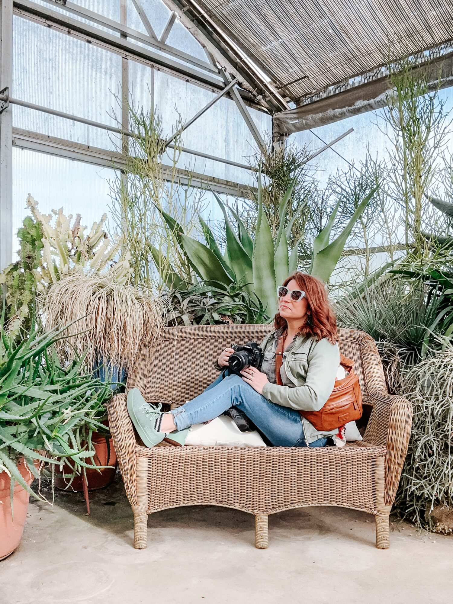Woman with sunglasses and a camera sitting on a wicker sofa in a greenhouse with plants and glass walls.