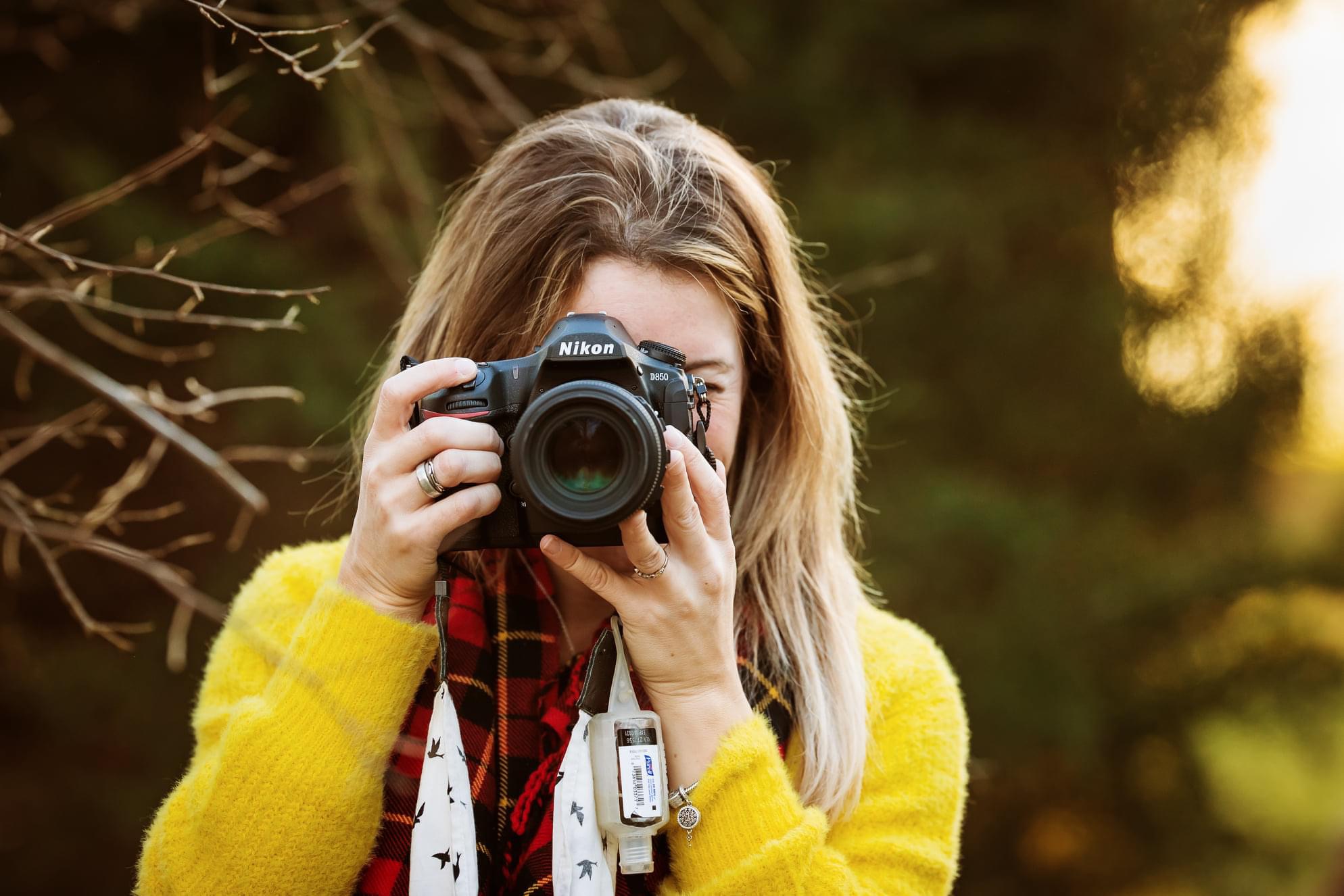 Person with long hair holding a camera up to their face outdoors, wearing a yellow sweater, sunlight in background.