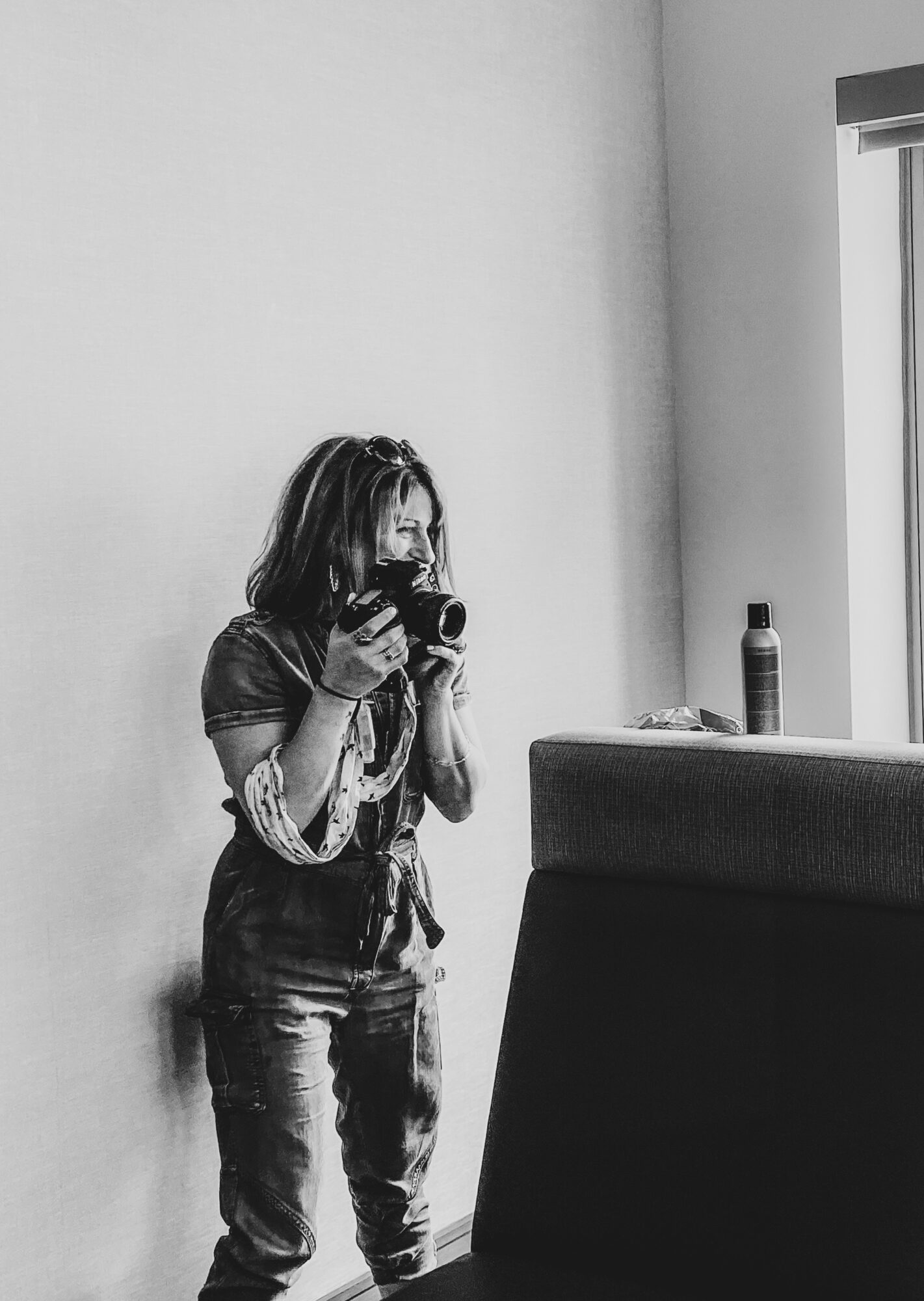 Person with long hair taking a photo with a camera, standing near a wall and window, in black and white.