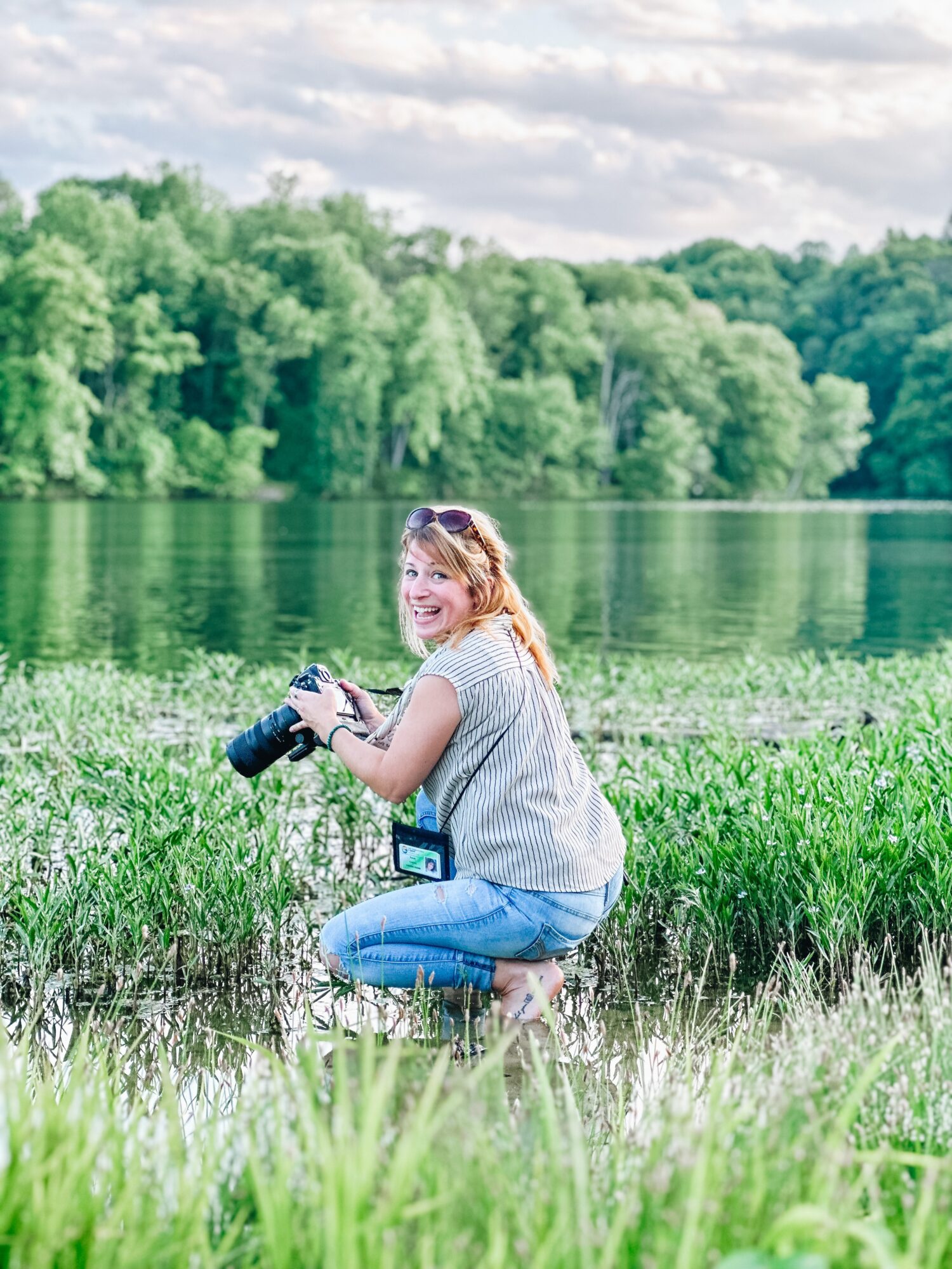 Woman crouching by a lake holding a camera, surrounded by green grass and trees, with a cloudy sky.