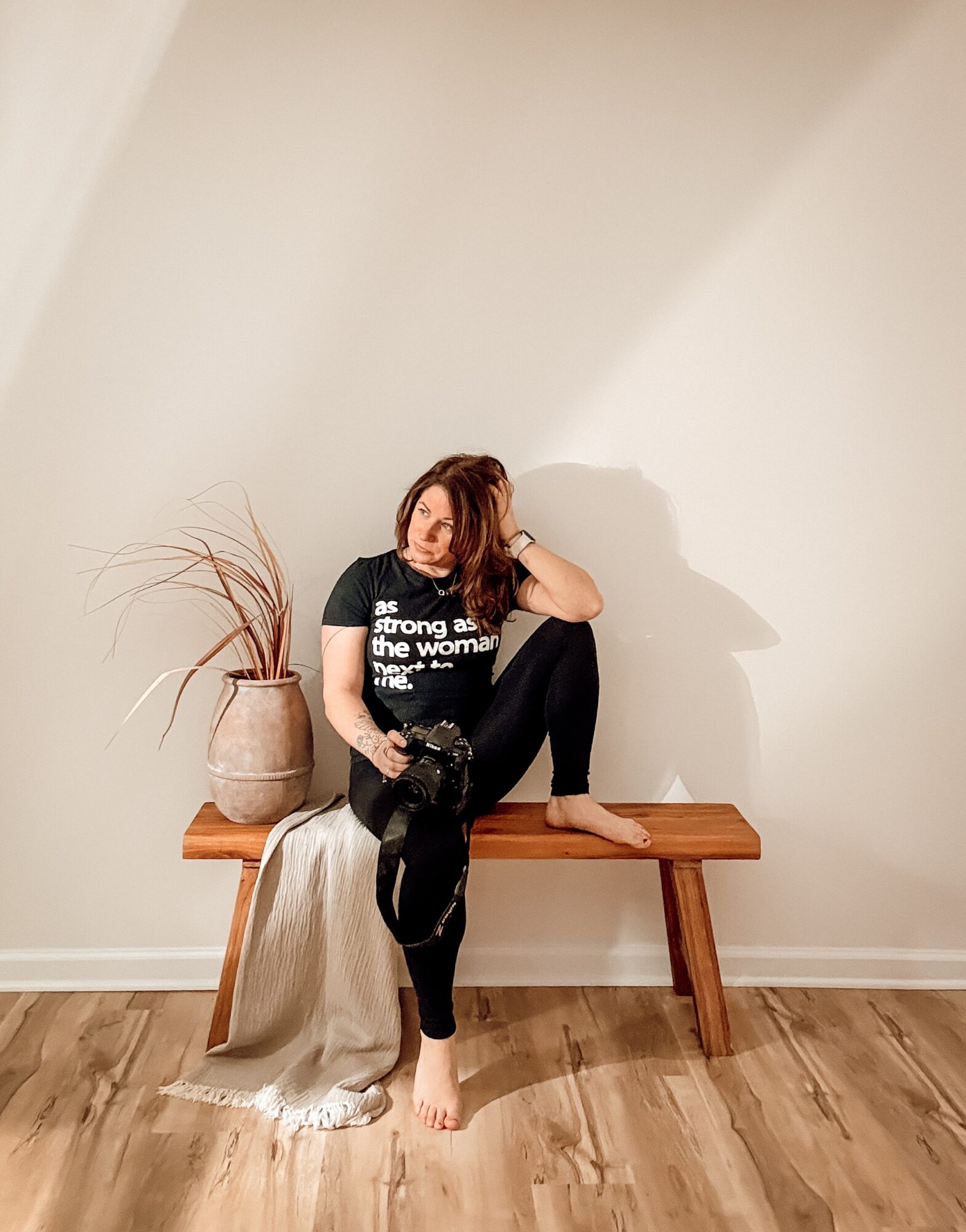 Woman sitting on a wooden bench with a plant in a vase beside her, leaning her head on her hand, barefoot, in a room with plain walls.