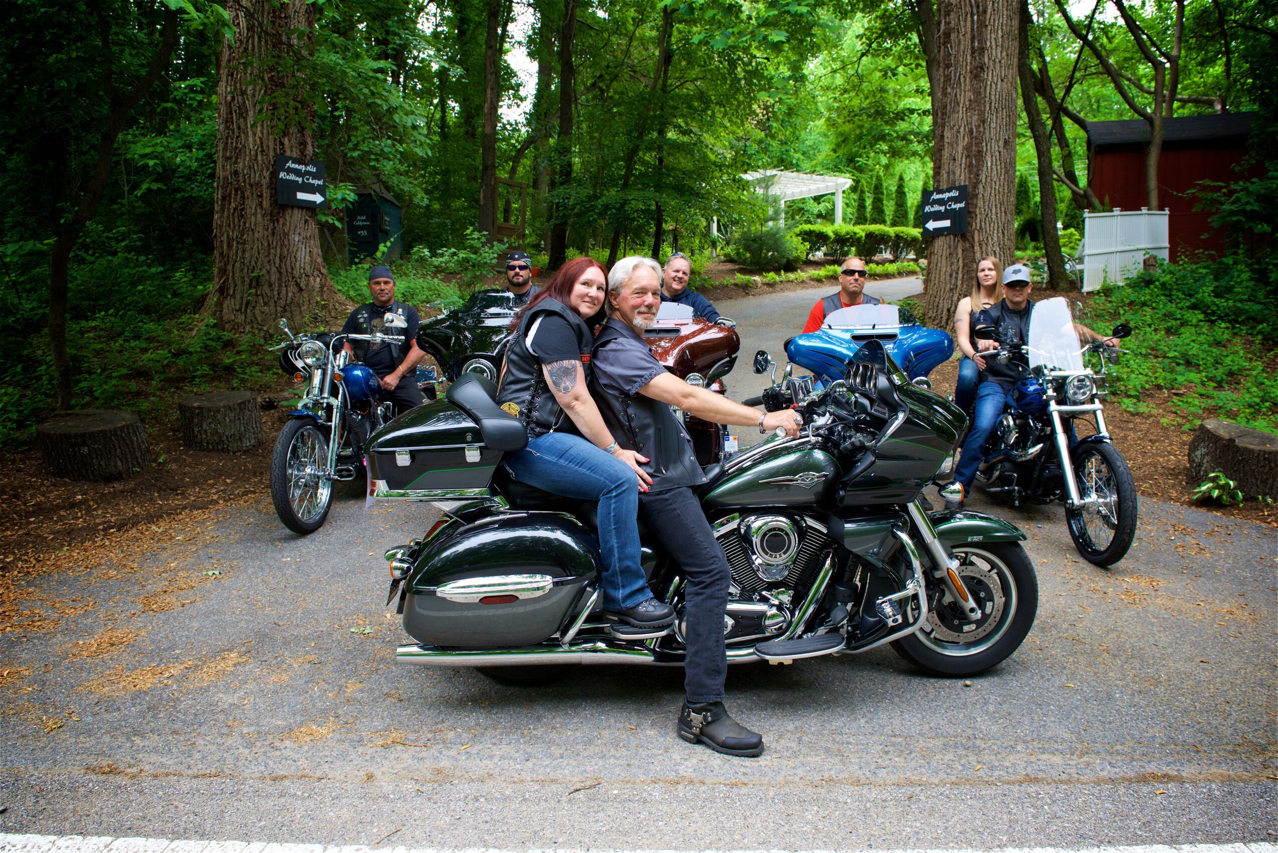 Group of people on motorcycles and a scooter in a wooded area with trees and signs, some smiling and looking at the camera.