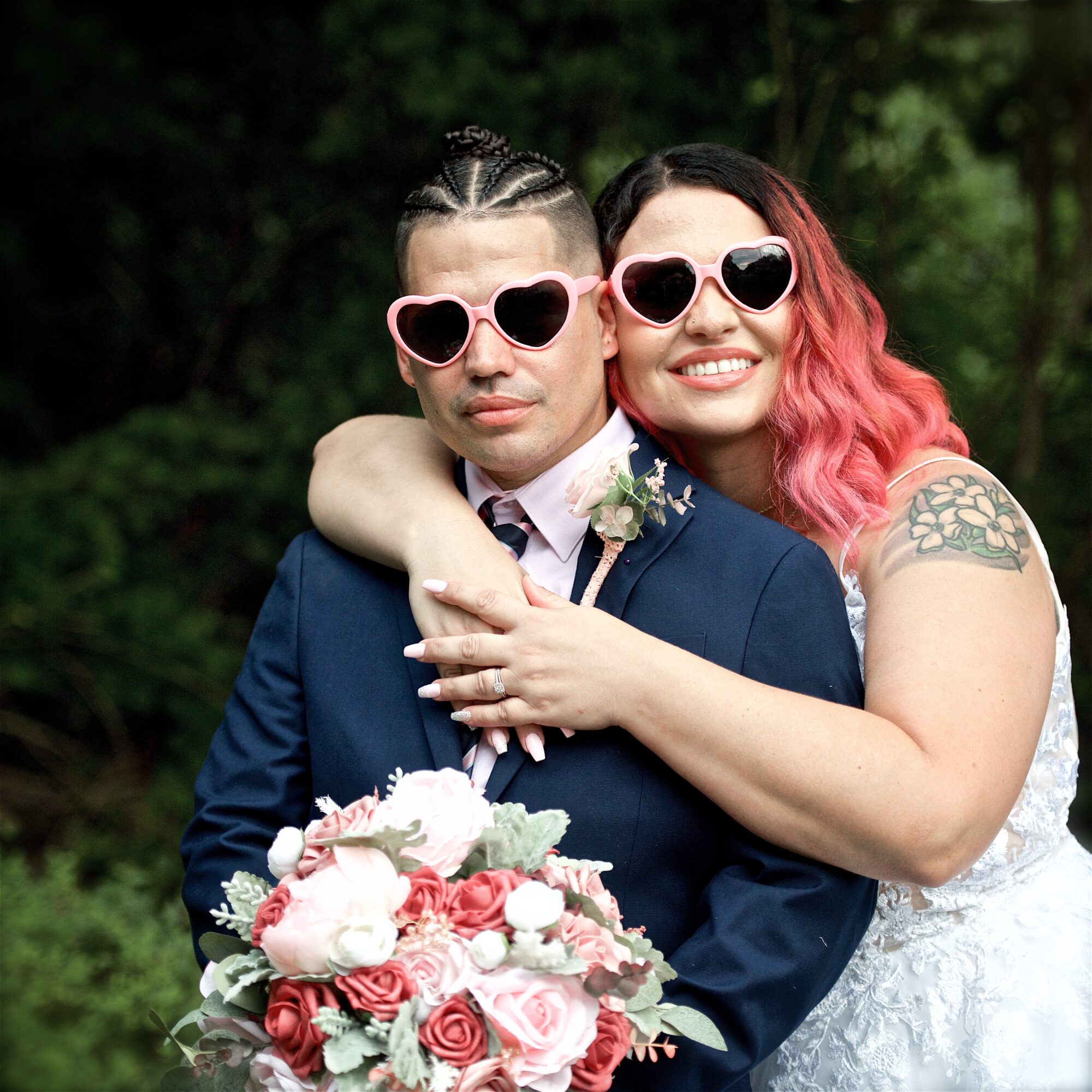 Two people, one with pink hair and sunglasses, hugging outdoors, holding a bouquet of pink and white flowers.