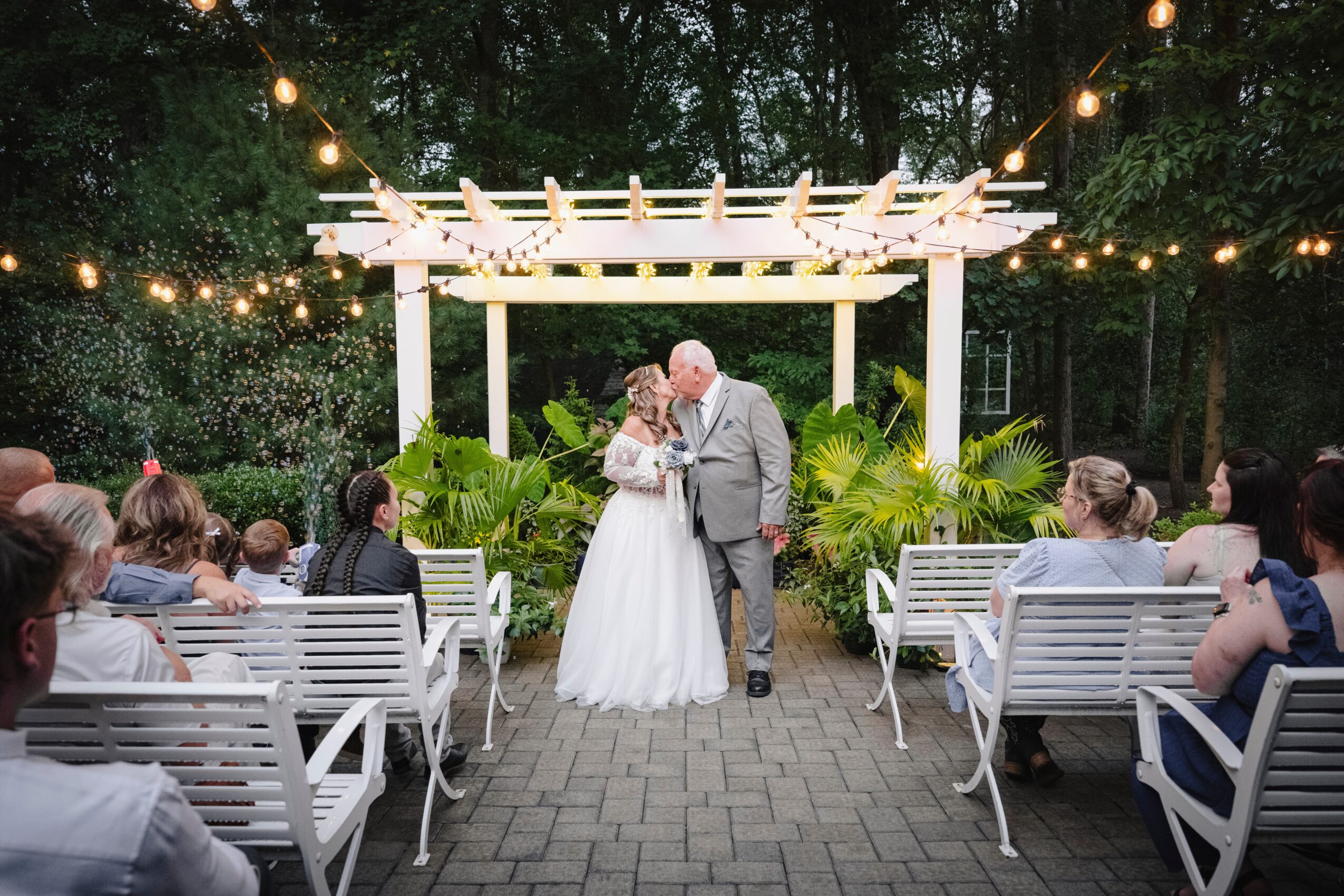 Bride and groom kiss under a white pergola at an outdoor wedding ceremony, guests seated on benches, string lights overhead.