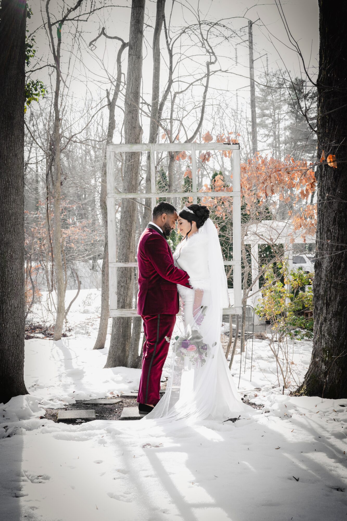 A couple in wedding attire stands close together in a snowy forest, holding hands and looking at each other.