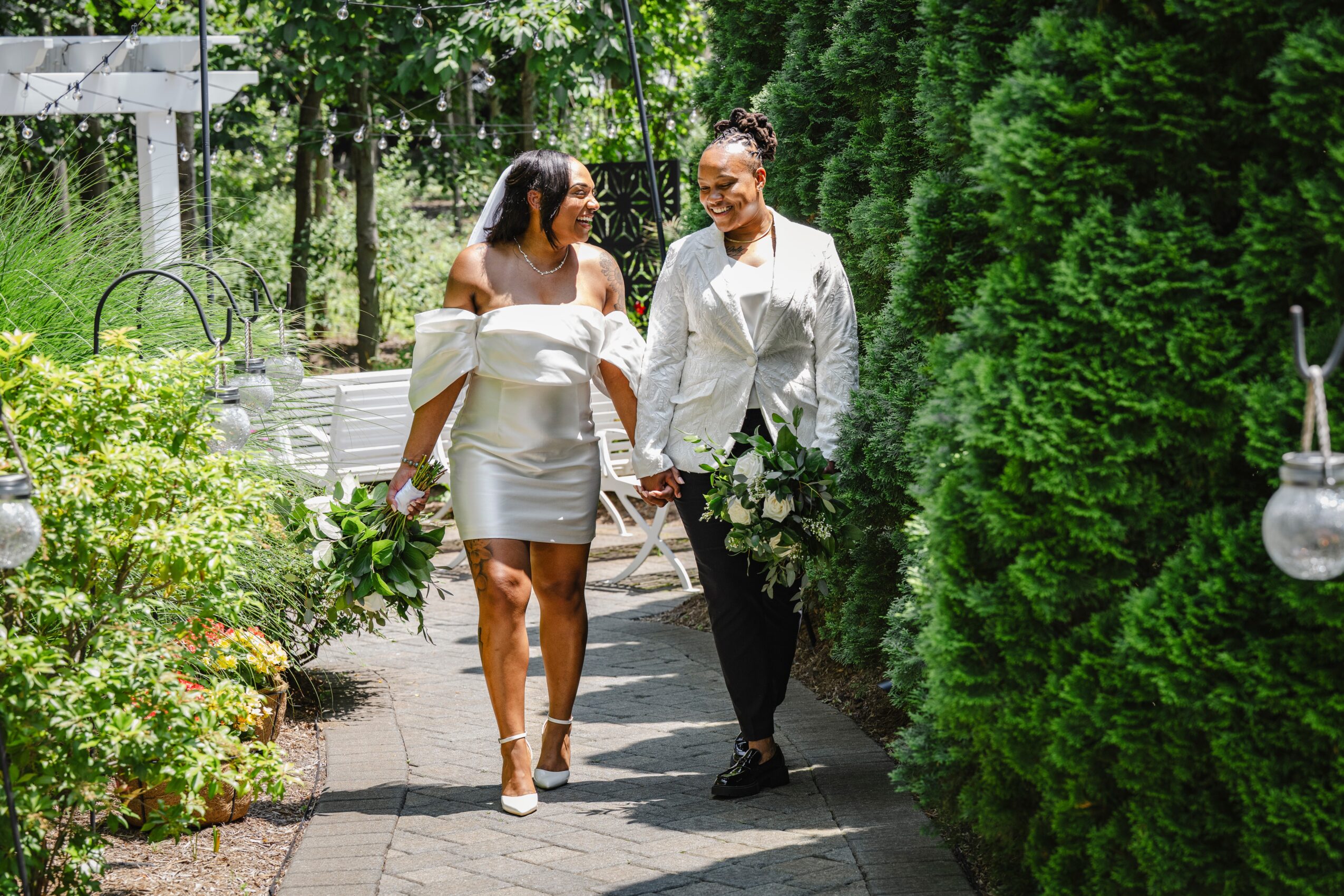Two women walk hand in hand along a garden path surrounded by greenery and flowers, smiling at each other.