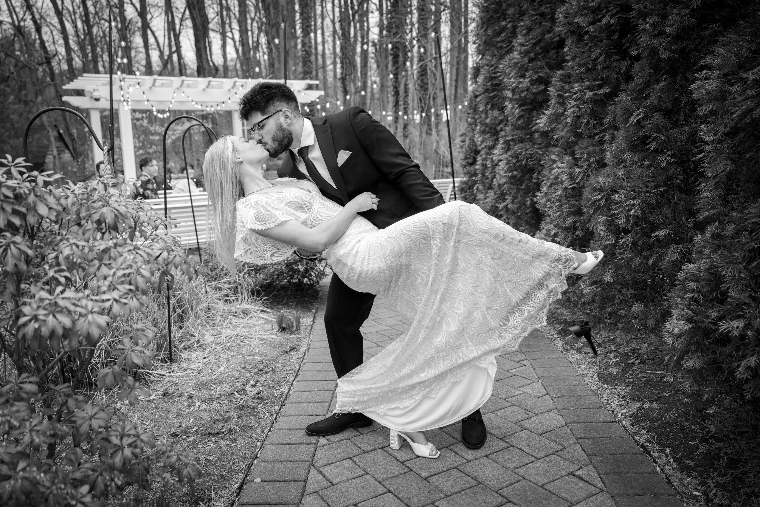 A couple dancing outdoors on a paved path, surrounded by trees and bushes, with a gazebo in the background.