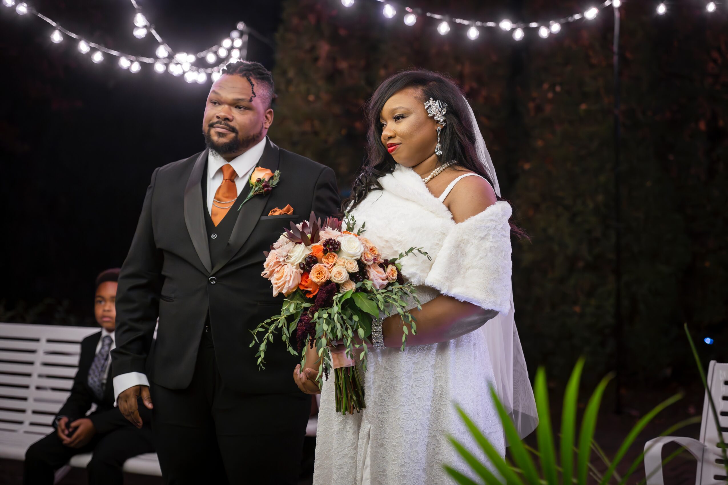 A bride and groom stand outdoors at night, with string lights overhead, the bride holding a bouquet, the groom smiling.