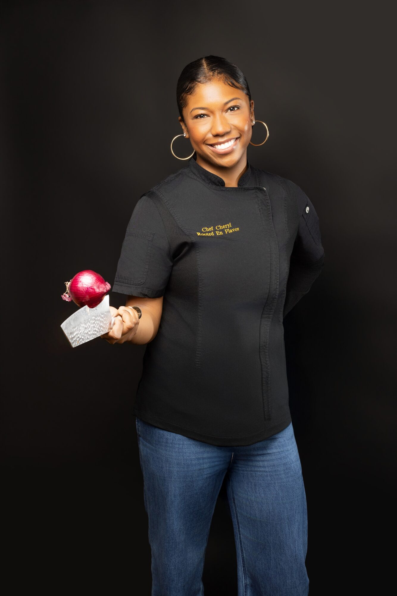 Smiling woman in black chef's coat holding a dessert with pink topping, against black background.