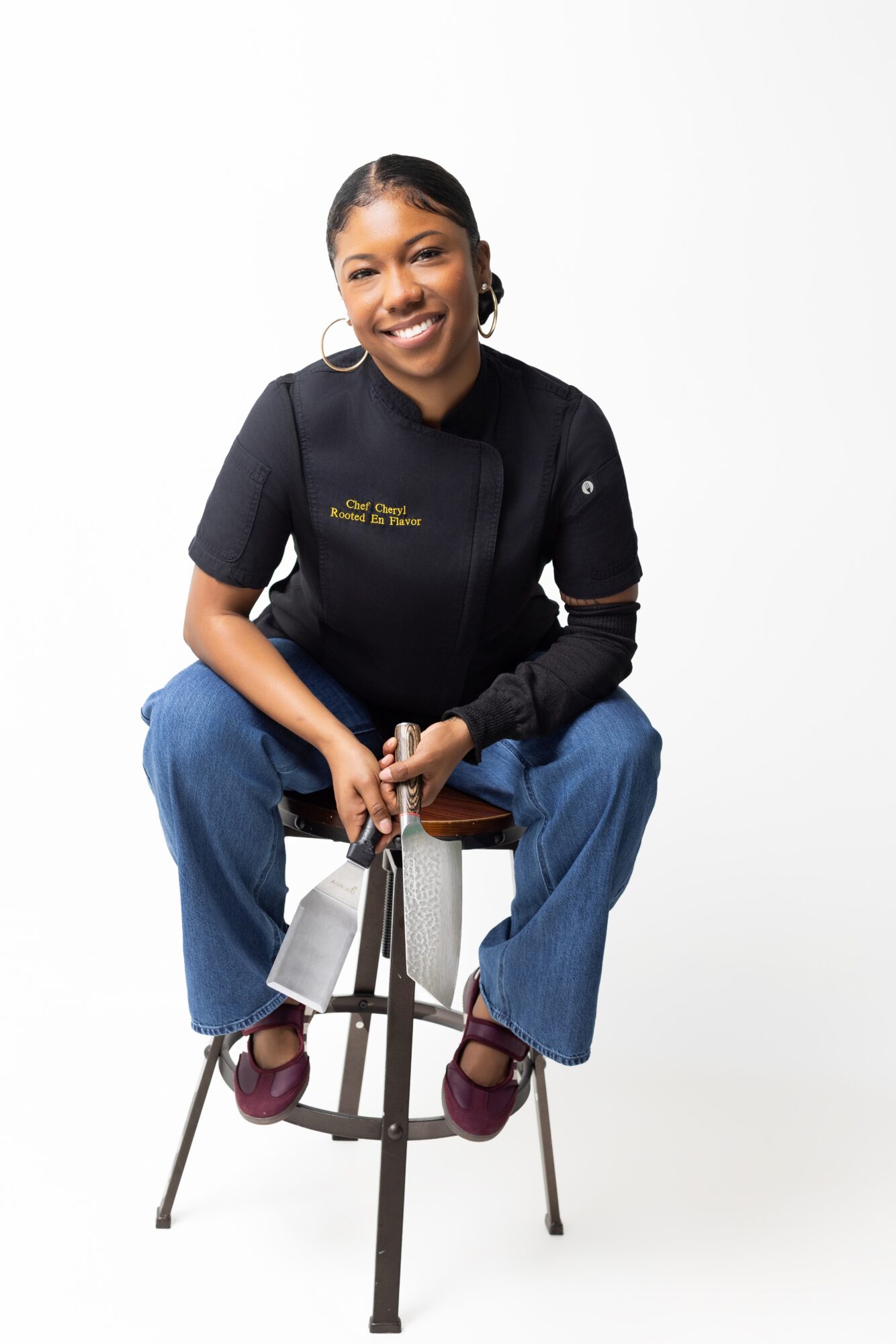 Young woman sitting on a stool, smiling, wearing a black top and blue jeans, against a white background.