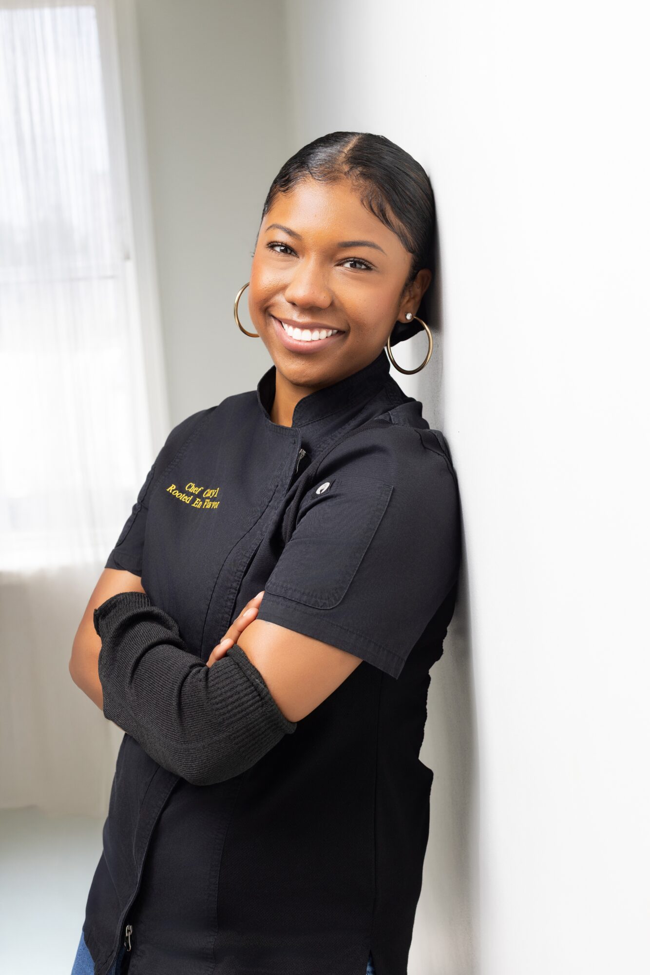 Smiling woman with arms crossed, wearing a black uniform, standing indoors near a window with curtains.
