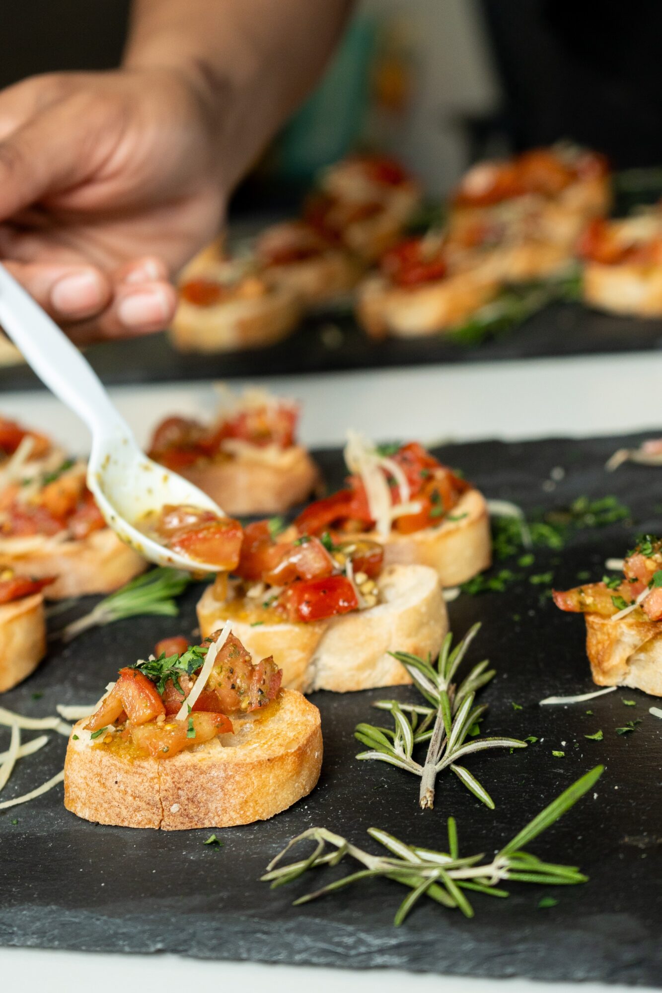Person adding sauce to small bread slices topped with vegetables on a black slate surface, garnished with rosemary sprigs.