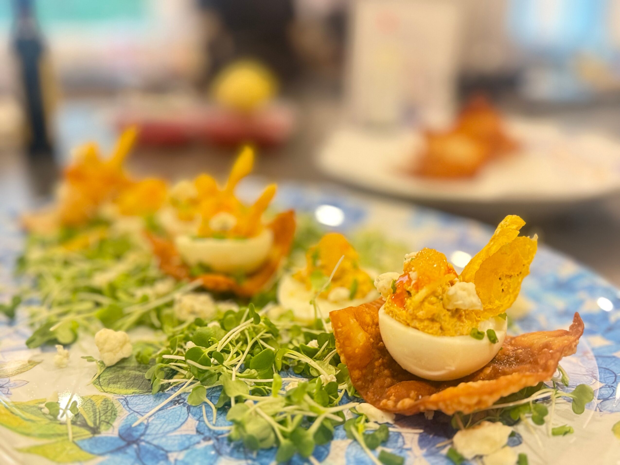 Deviled eggs garnished with chips and herbs on a bed of greens, with blurred background of a dining setting.
