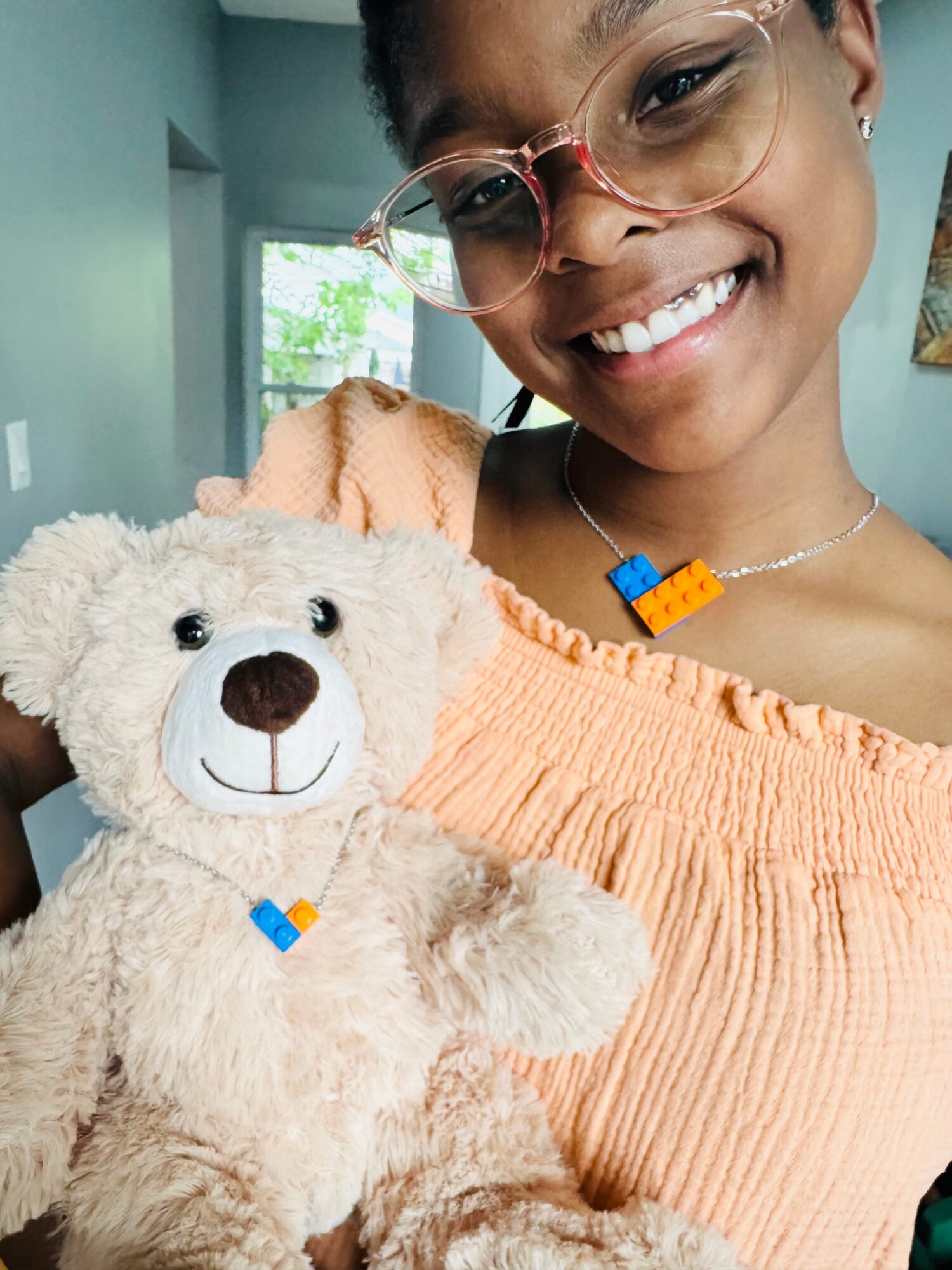 Smiling person with glasses holding a teddy bear, wearing an orange top, in a room with a window and artwork.