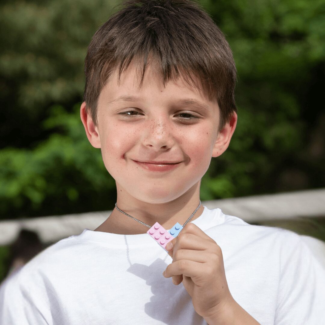 Boy smiling outdoors holding a small colorful object, background of green trees and bushes, wearing a white shirt.
