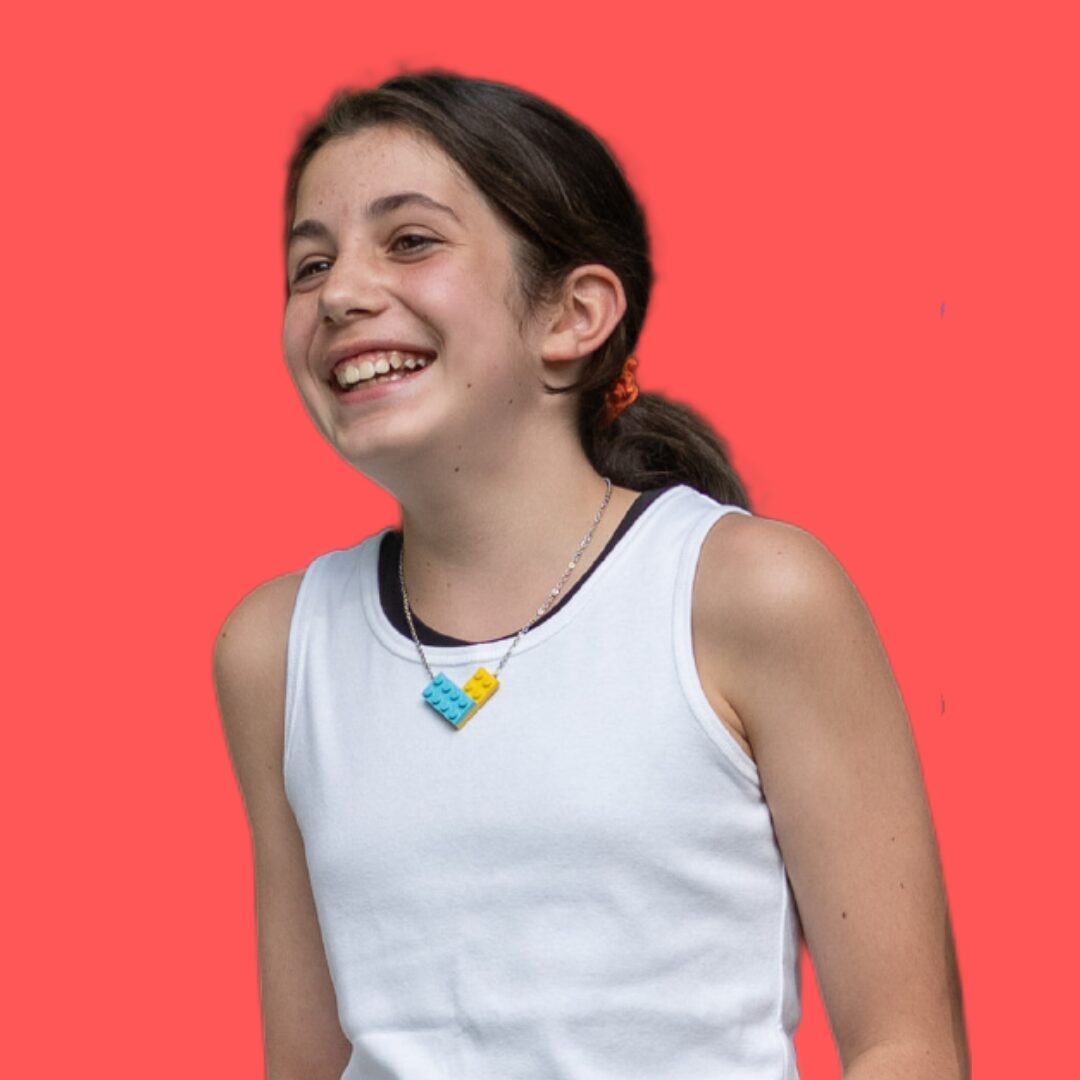 Smiling girl with dark hair in a ponytail, wearing a white sleeveless top and a colorful necklace, against a red background.
