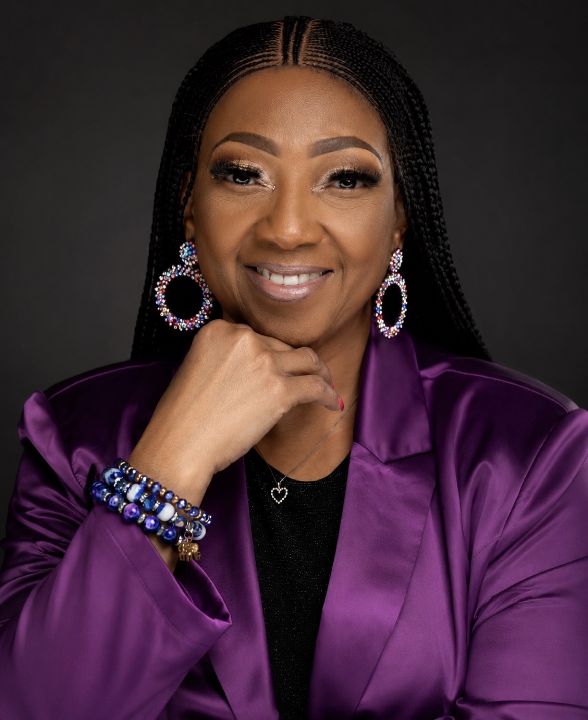 Portrait of a woman with braided hair, wearing large earrings, a purple satin jacket, and a black top, smiling.