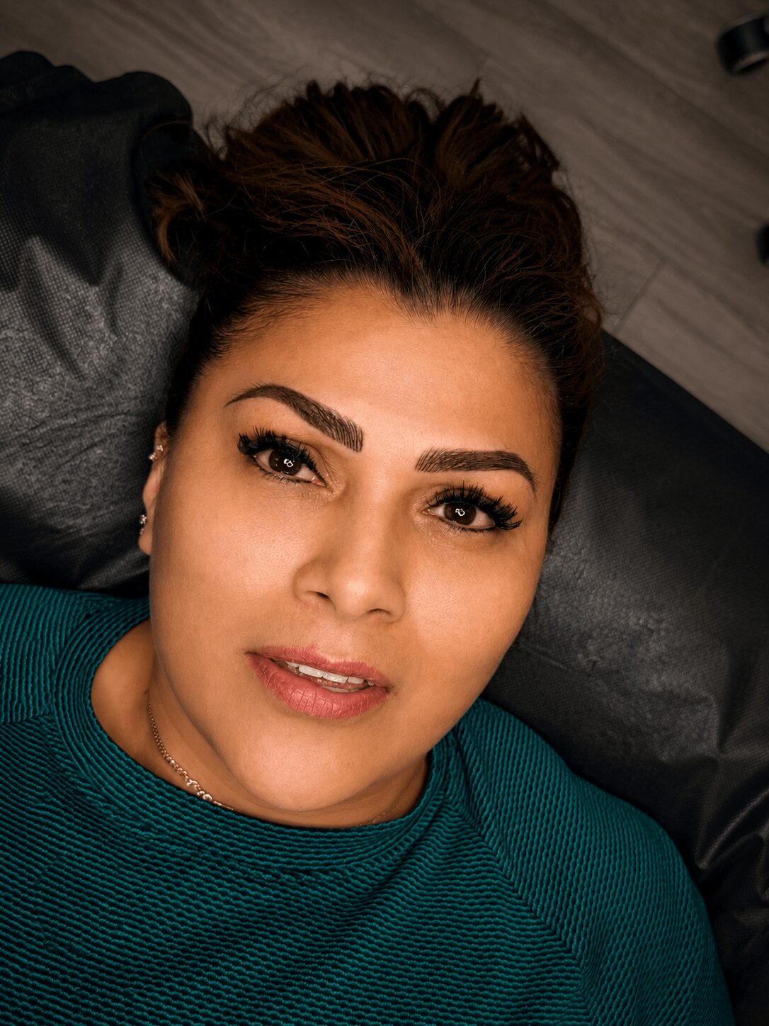 Woman with dark hair lying down, looking at the camera, wearing a teal top, with a neutral background.