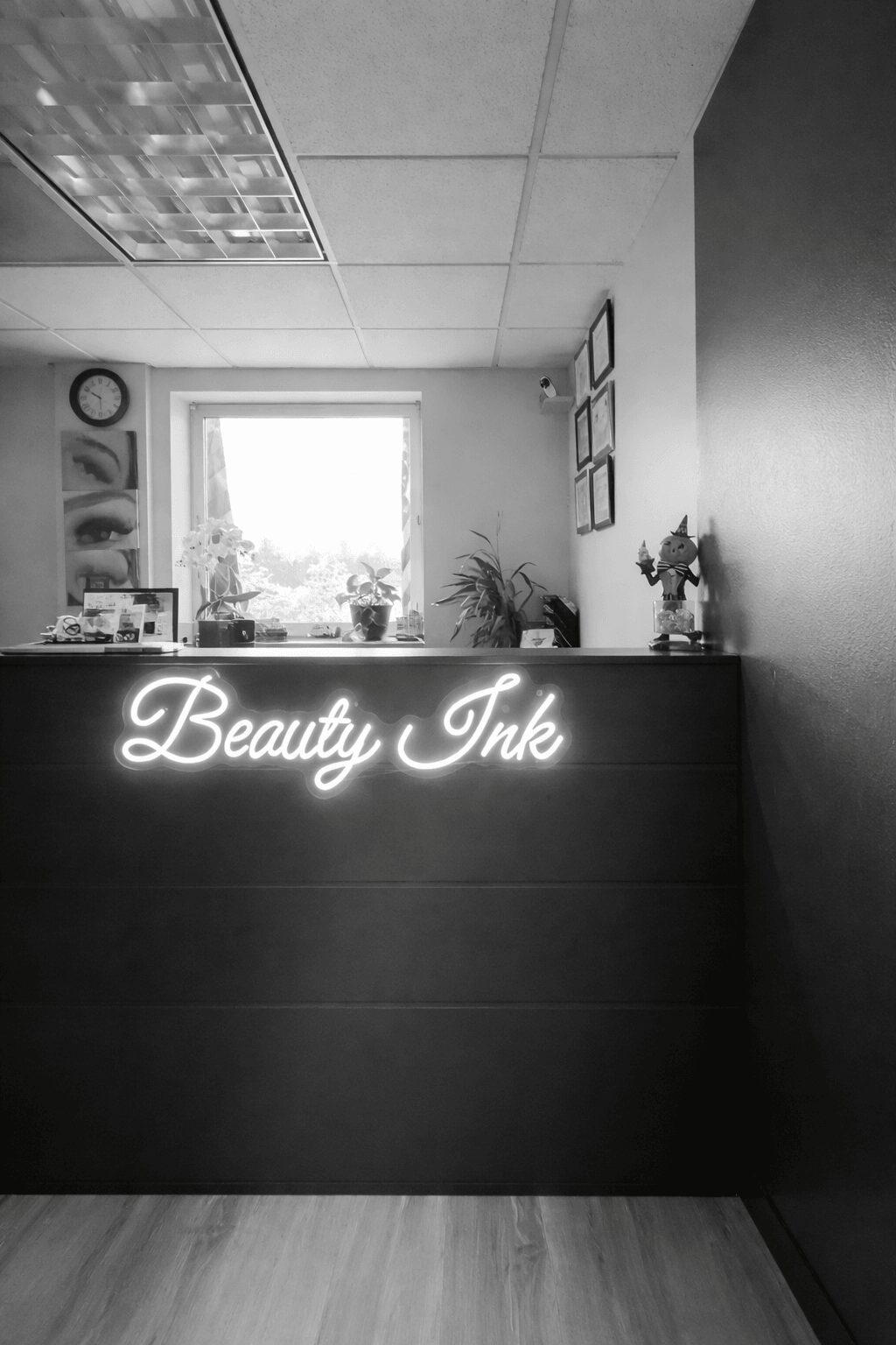 Interior space with a neon sign reading 'Beauty Ink' on a dark counter, window with plants, framed pictures, and ceiling tiles.