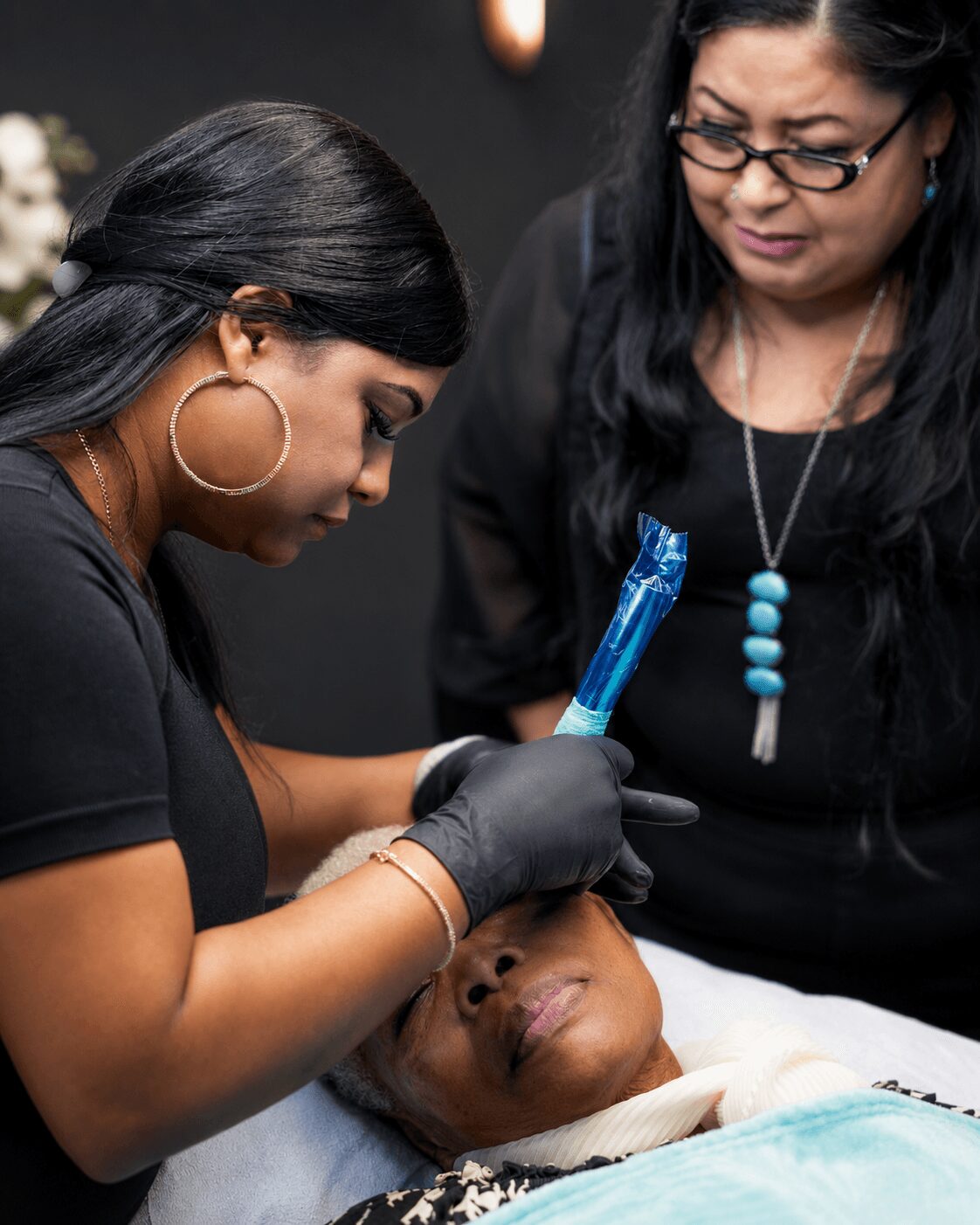 A woman in black gloves performs a procedure on a patient lying in a medical setting, with another woman observing closely.