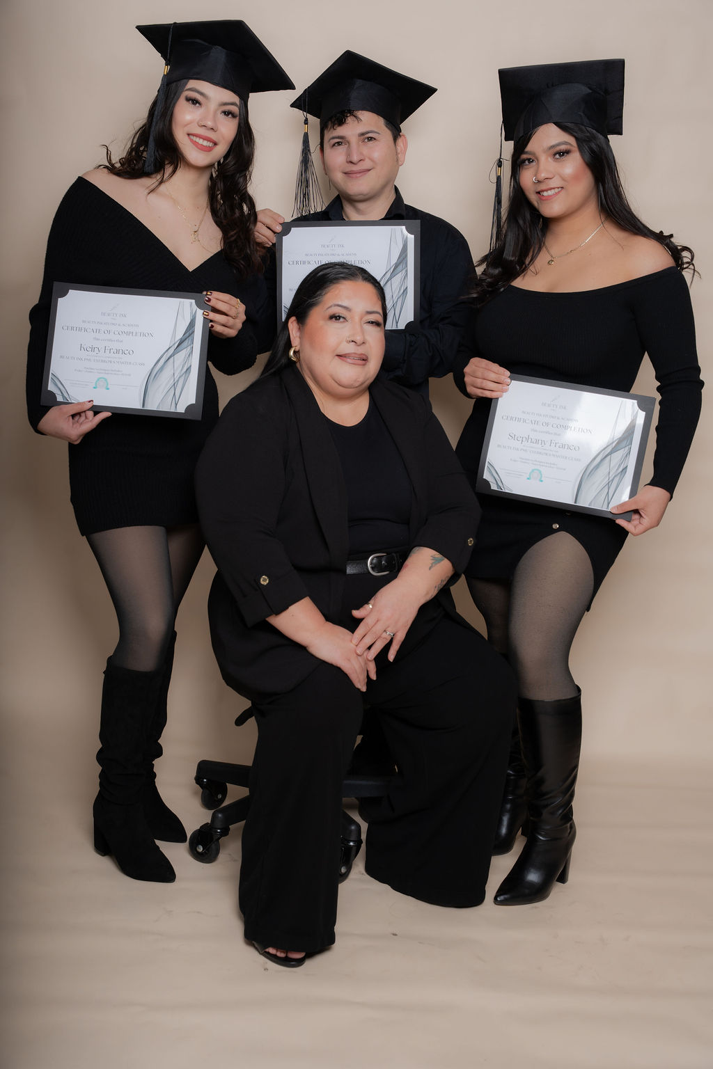 Four women, three standing and one sitting, holding diplomas, wearing black outfits and graduation caps, smiling, against a plain background.