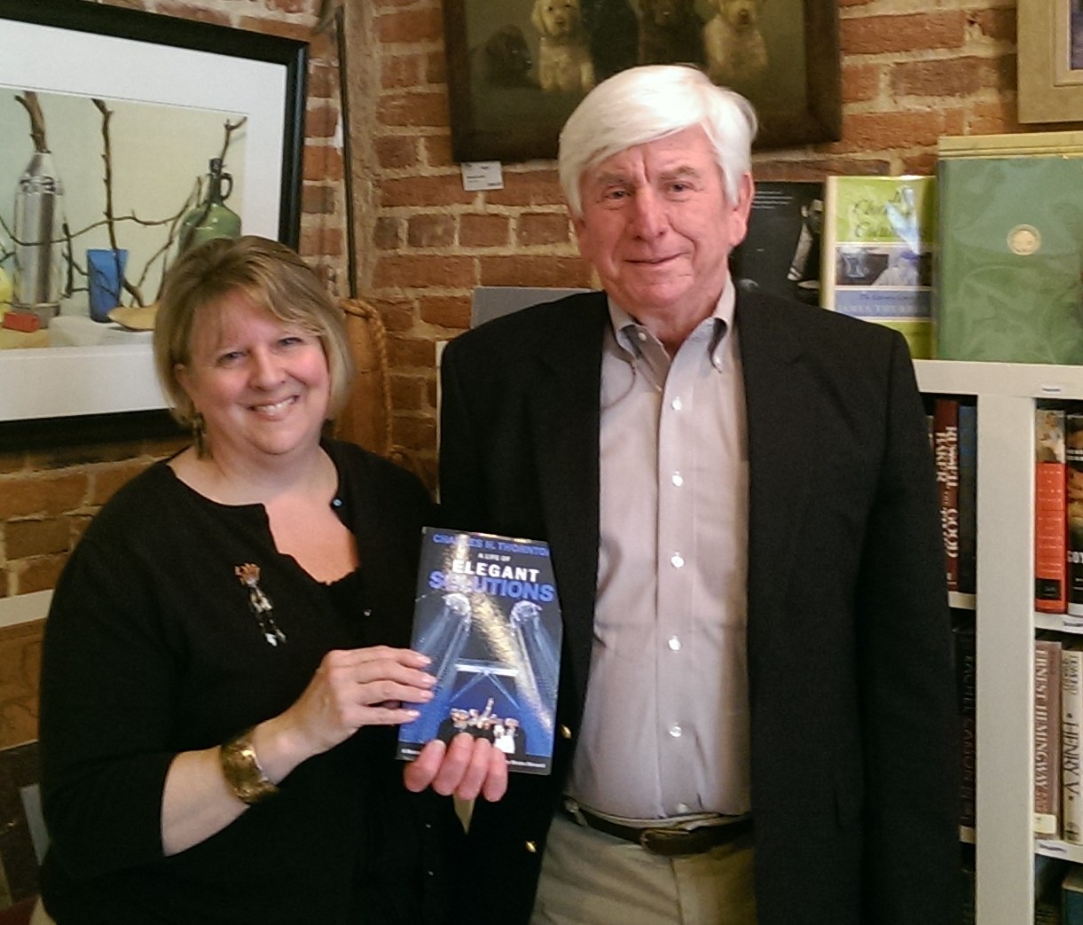 Two people standing indoors, holding a book, with artwork and bookshelves in the background.