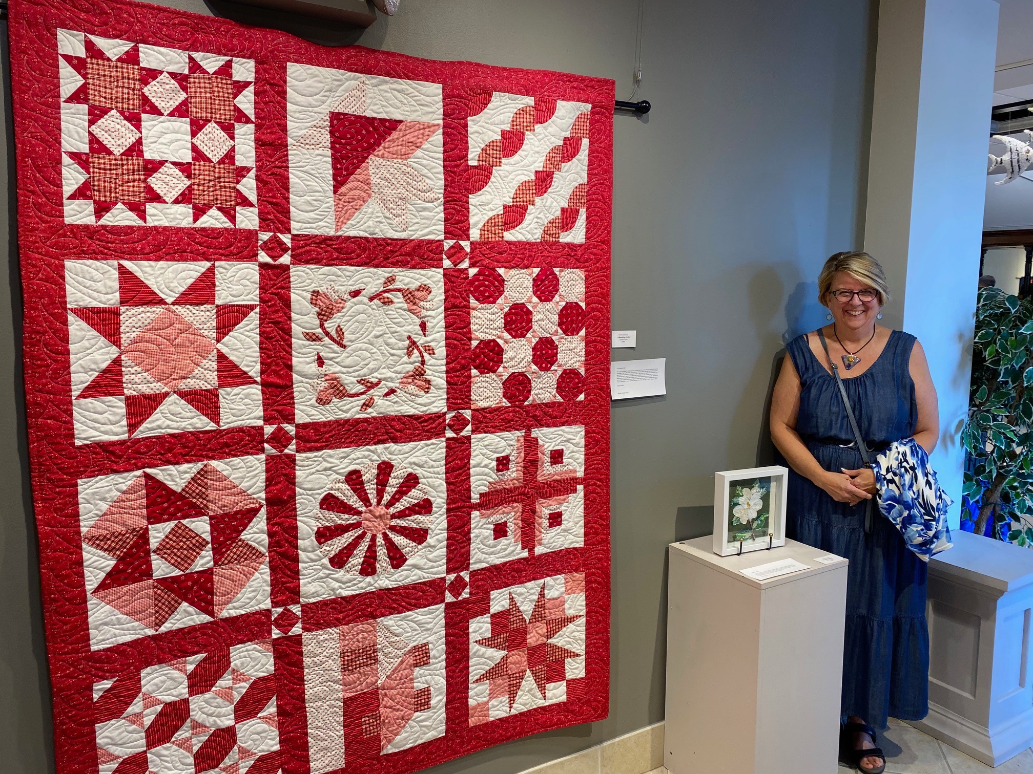 A woman in a blue dress and glasses stands next to a white pedestal with a framed picture, in front of a red and white quilt displayed on a wall.