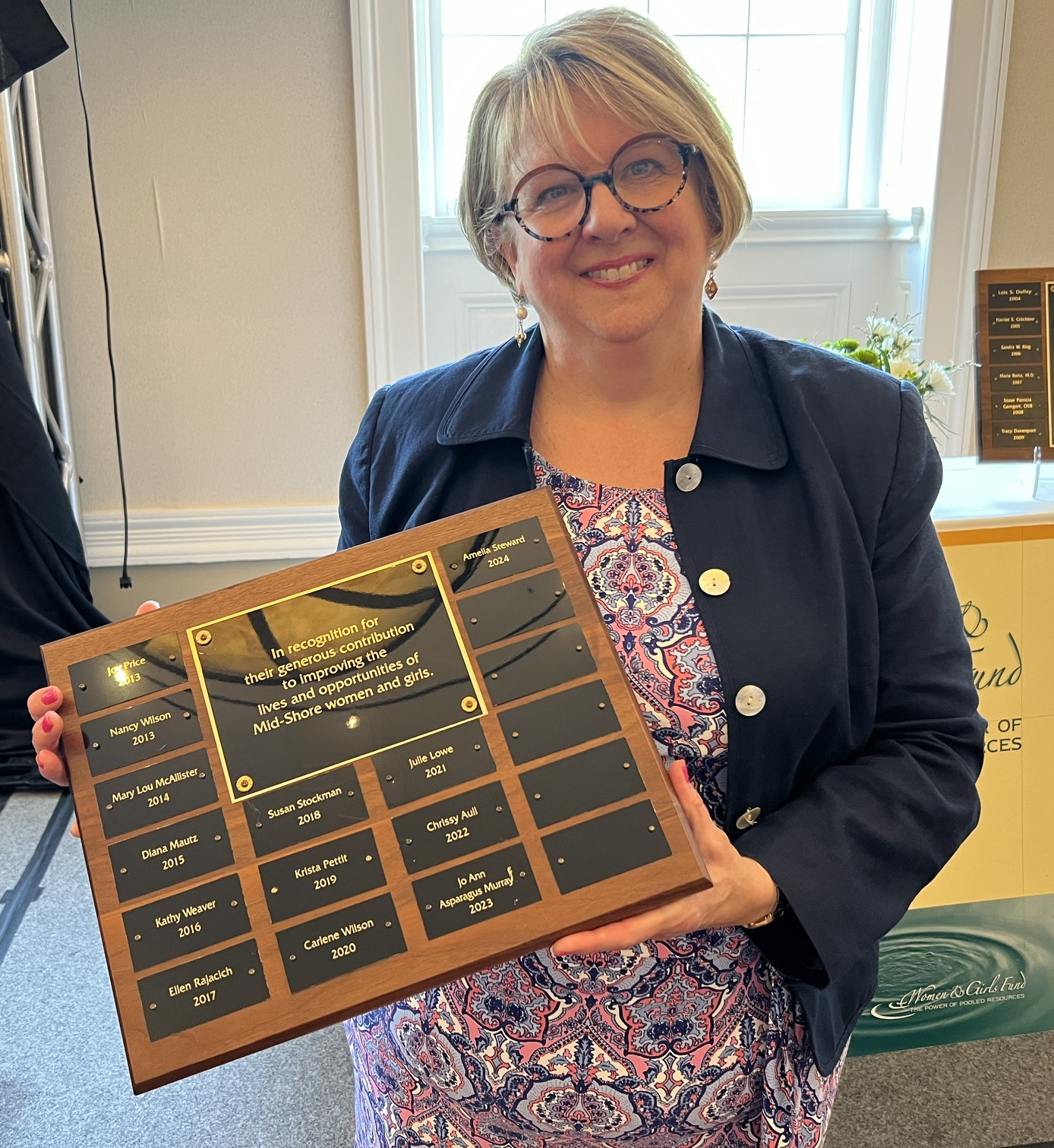 Woman with glasses and short hair holding a large award plaque, smiling indoors with a window behind her.