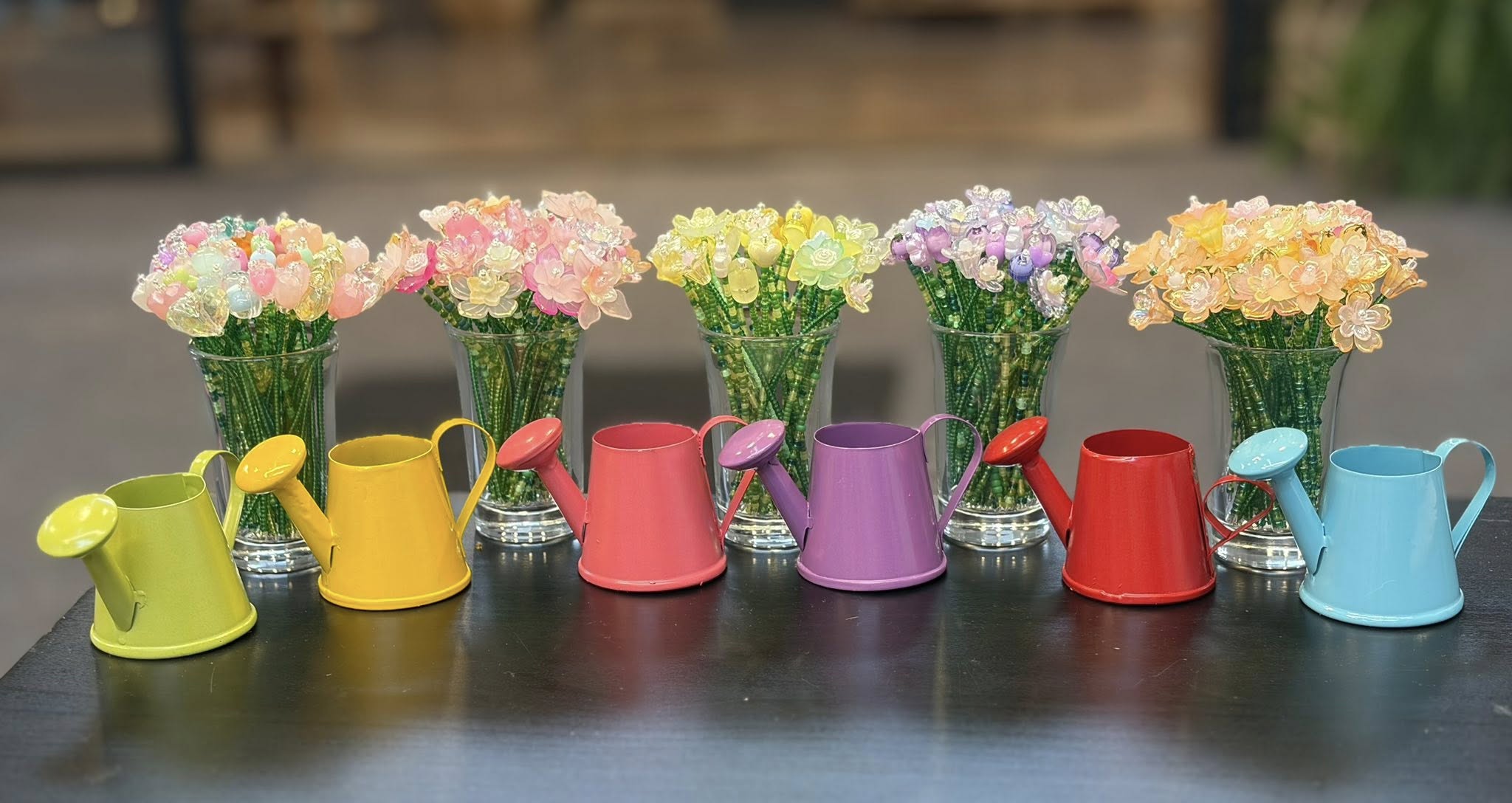Six colorful watering cans with flowers in glass vases arranged in a row on a dark surface.