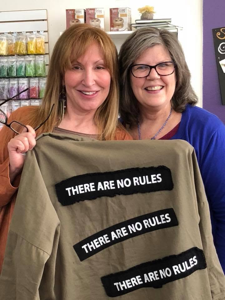 Two women smiling, holding a shirt with the text 'There are no rules' repeated three times, in a store setting.