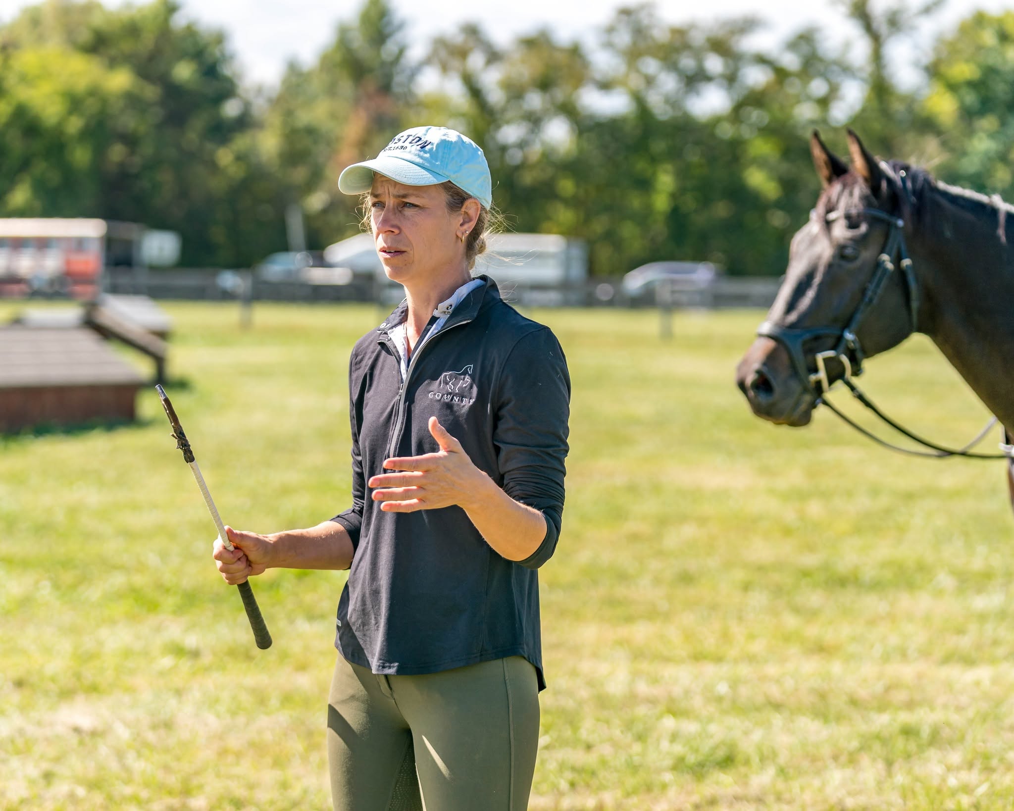 Woman holding a riding crop and talking to a horse outdoors on a grassy field.