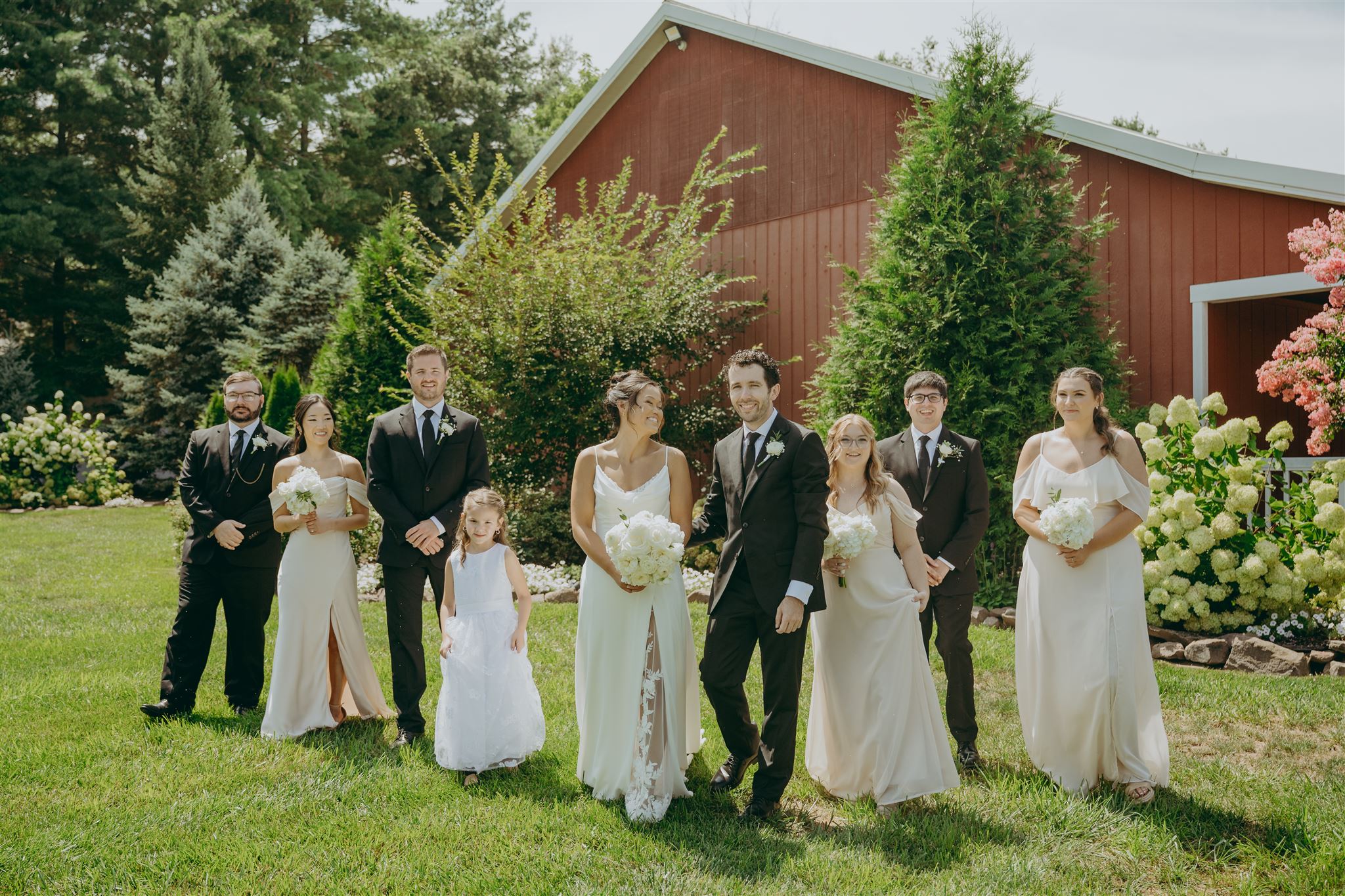 Group of people dressed in formal wedding attire standing on grass in front of a red barn and trees.