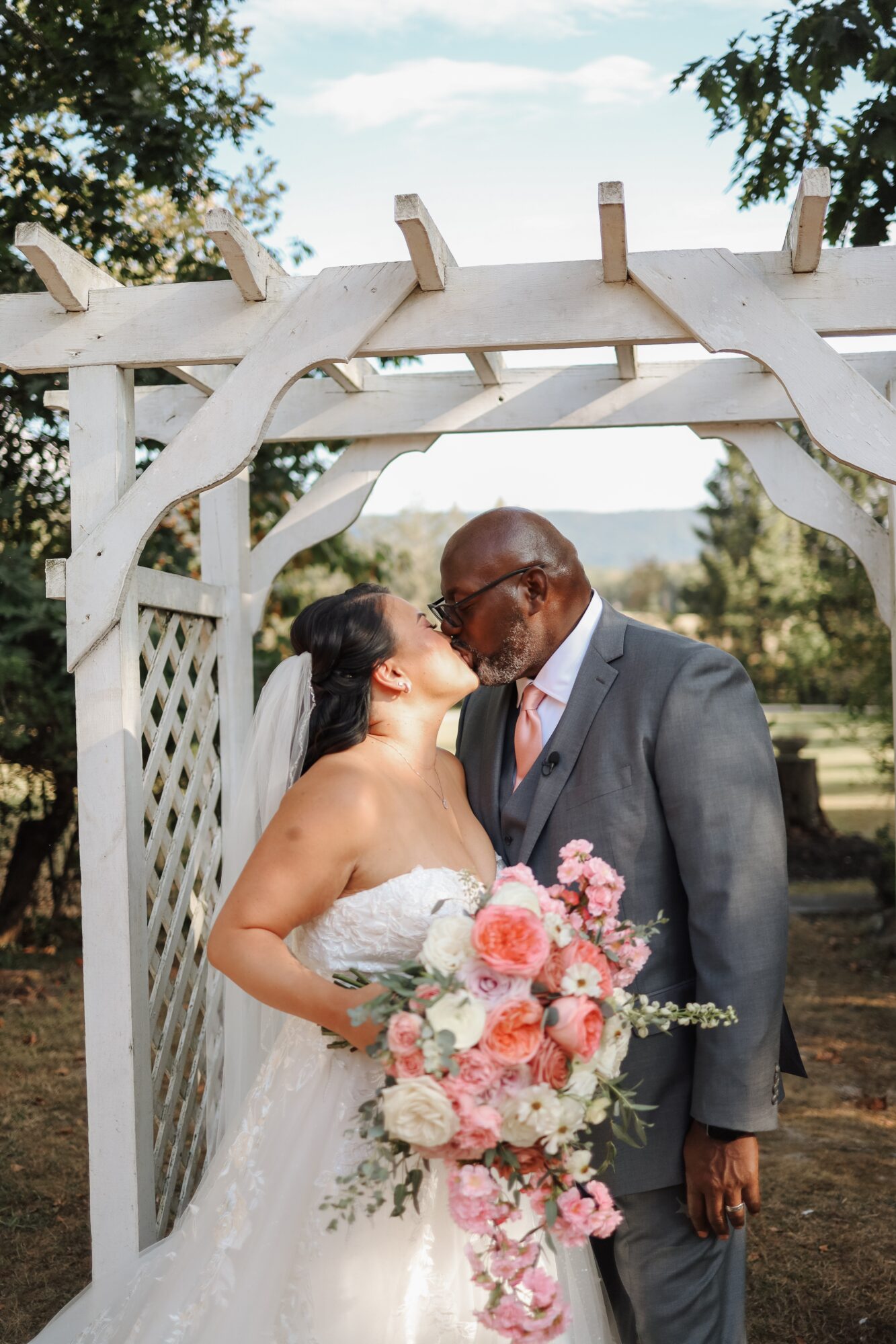 Bride and groom sharing a kiss under a white wooden arch outdoors, with trees and sky in the background.