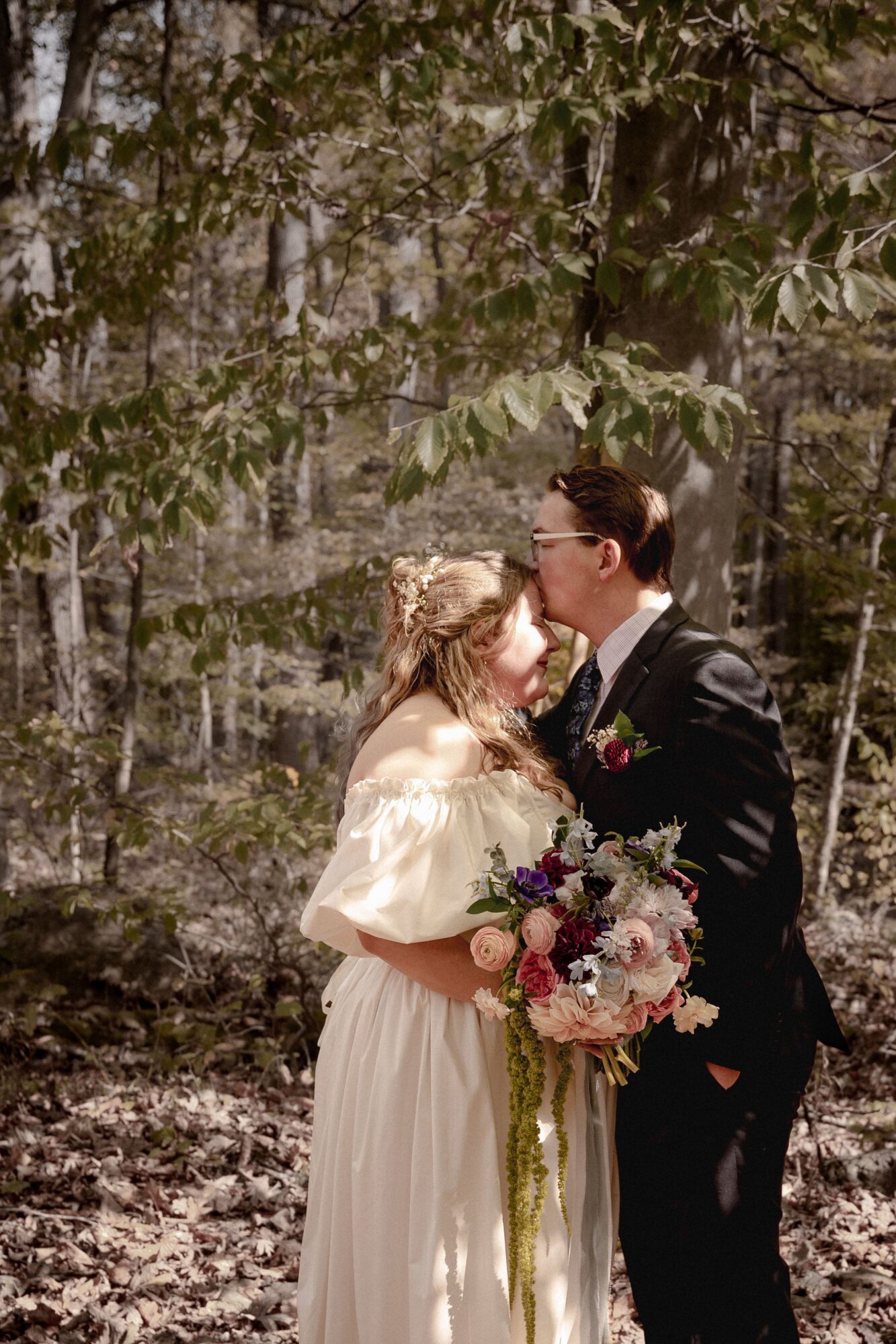 A couple in wedding attire standing outdoors among trees, holding a bouquet of flowers, facing each other closely.