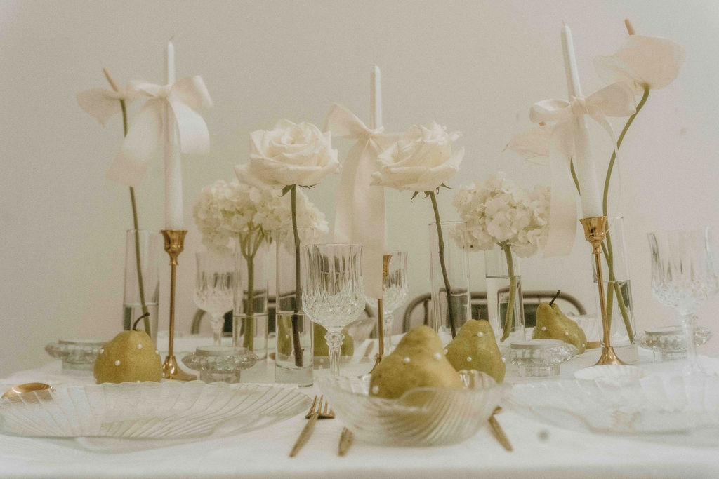 Table setting with white flowers, candles, and decorative items, arranged on a light-colored tablecloth.