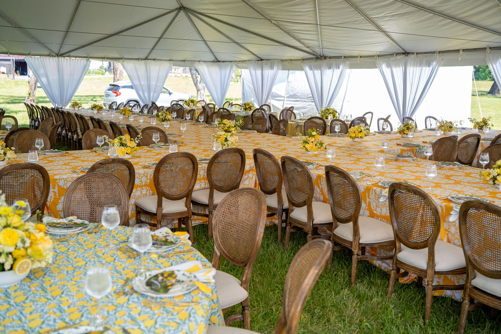 Large outdoor event tent with round tables, chairs, and yellow floral centerpieces, set for a meal, with grass visible outside.