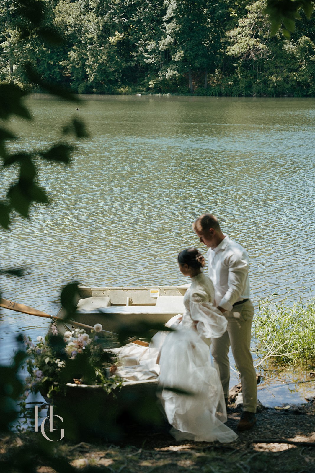 Two people stand near a lake with trees in the background, one in a white dress and the other in a white shirt and pants, by a boat.
