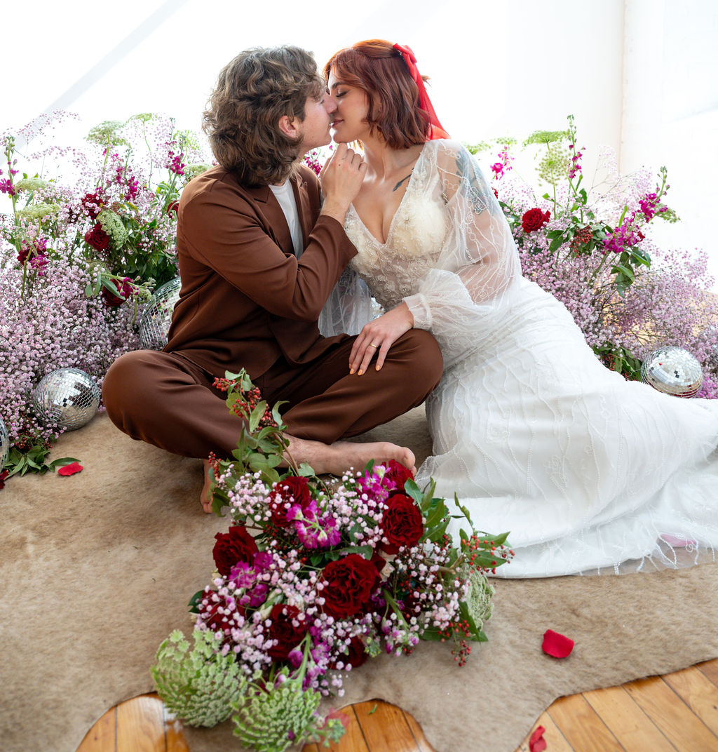 Couple sitting on floor, kissing, surrounded by flowers and a bouquet, in a bright setting with natural light.